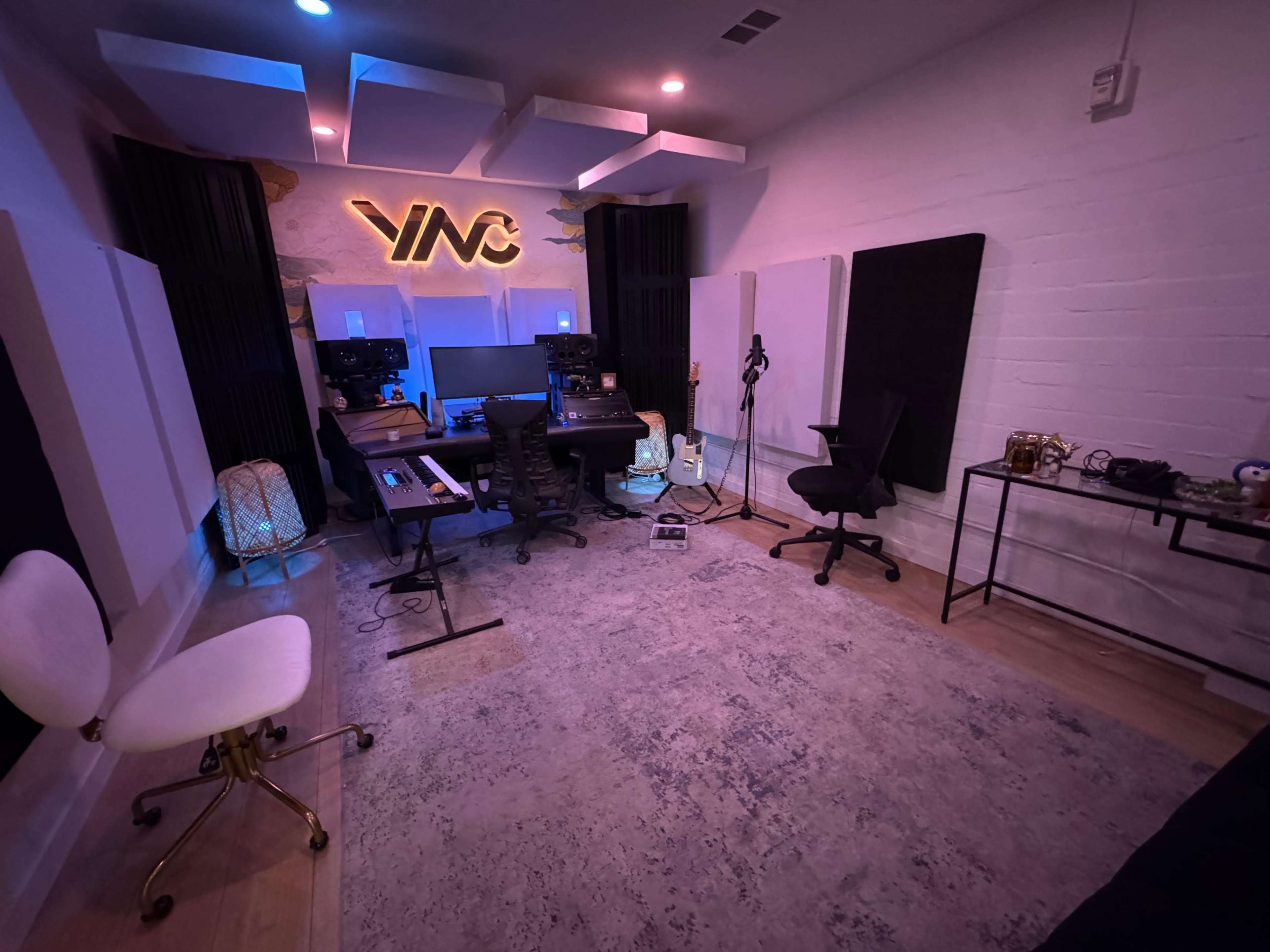 The image shows a music studio with a mixing desk, keyboard, guitar, microphones, and soundproofing panels on the walls.