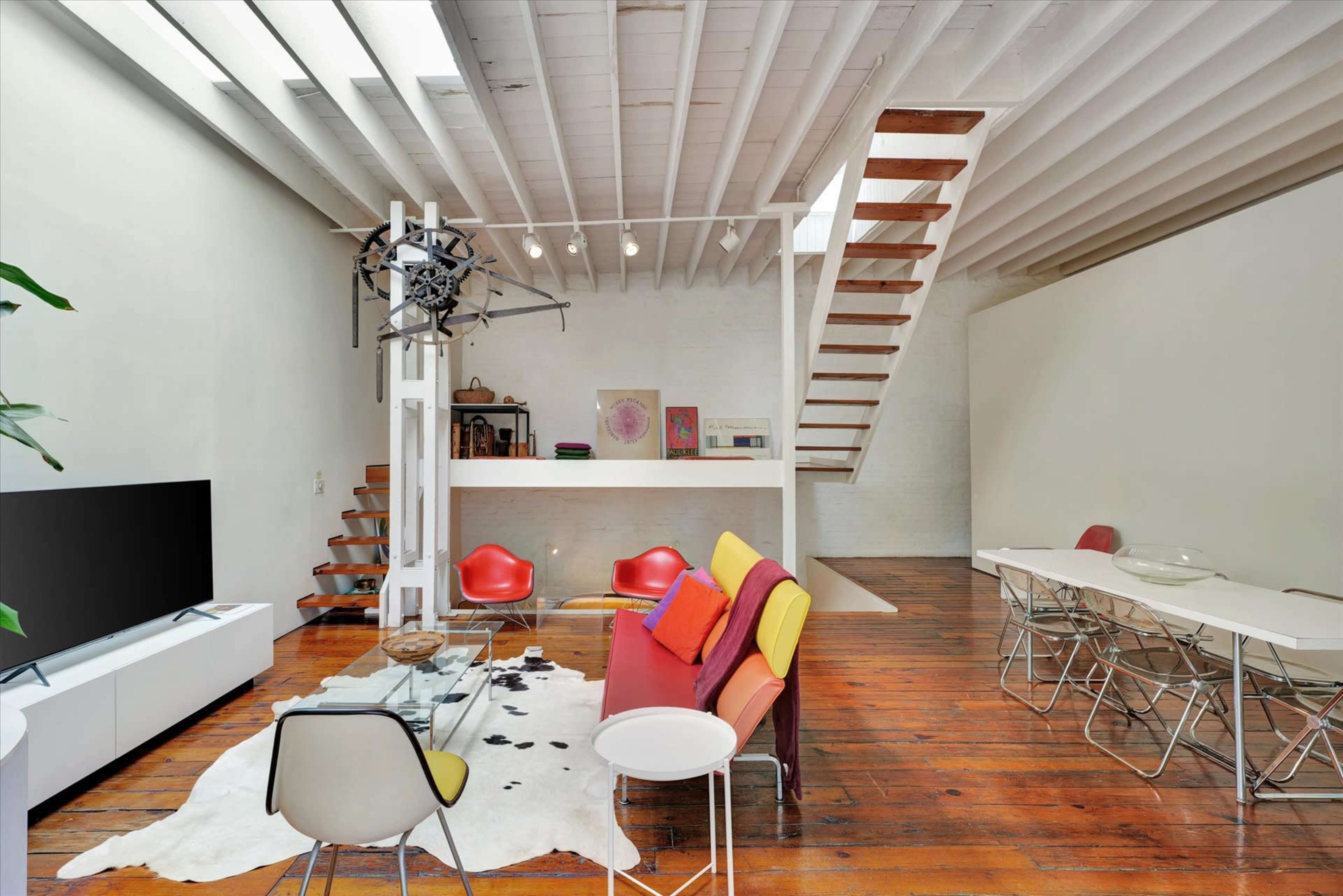 A modern living space with wooden floors, a staircase, a glass coffee table, and colorful seating arranged around a cowhide rug.