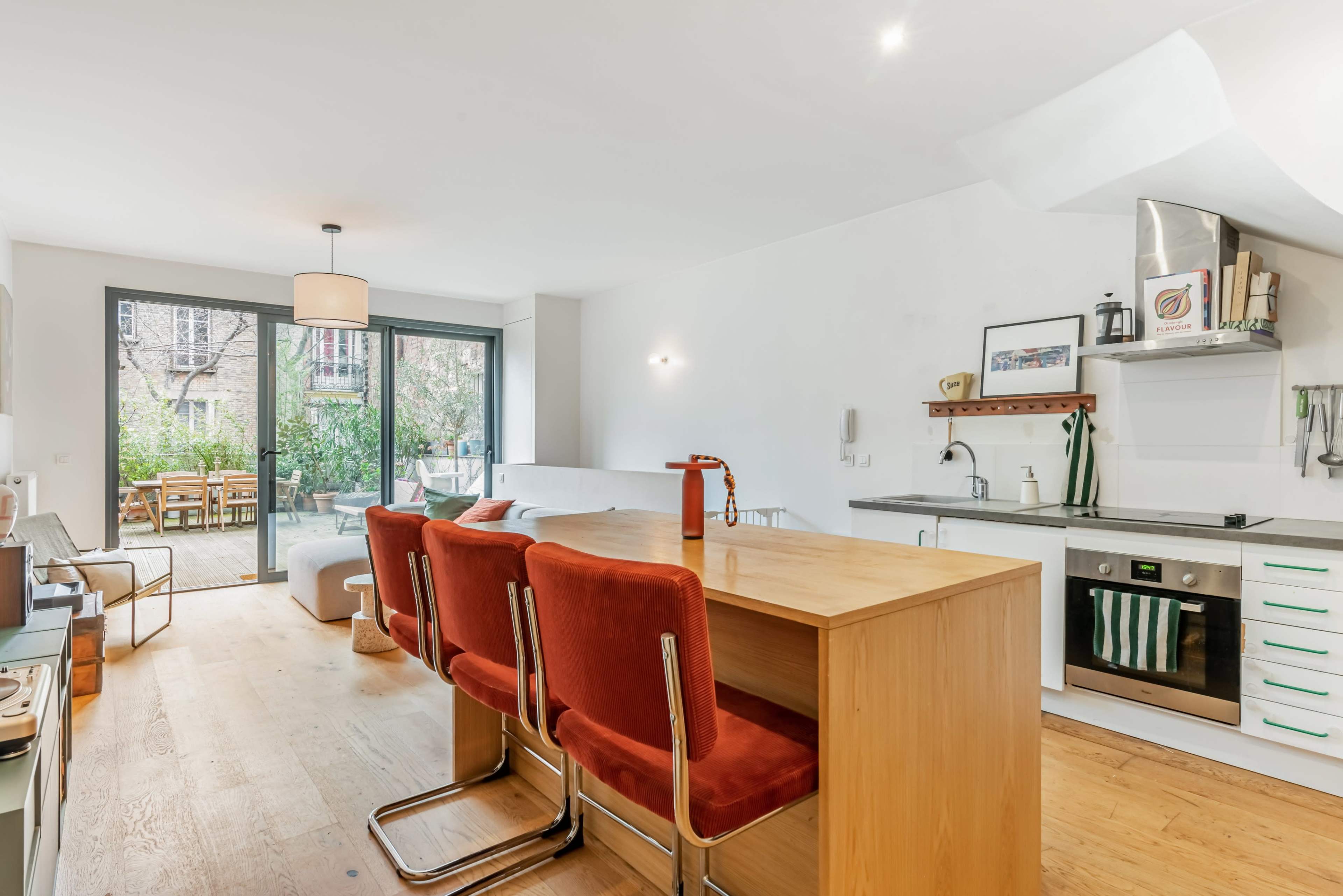 A modern kitchen and dining area featuring a wooden table with red chairs, an organized kitchen space, and large windows overlooking a garden.