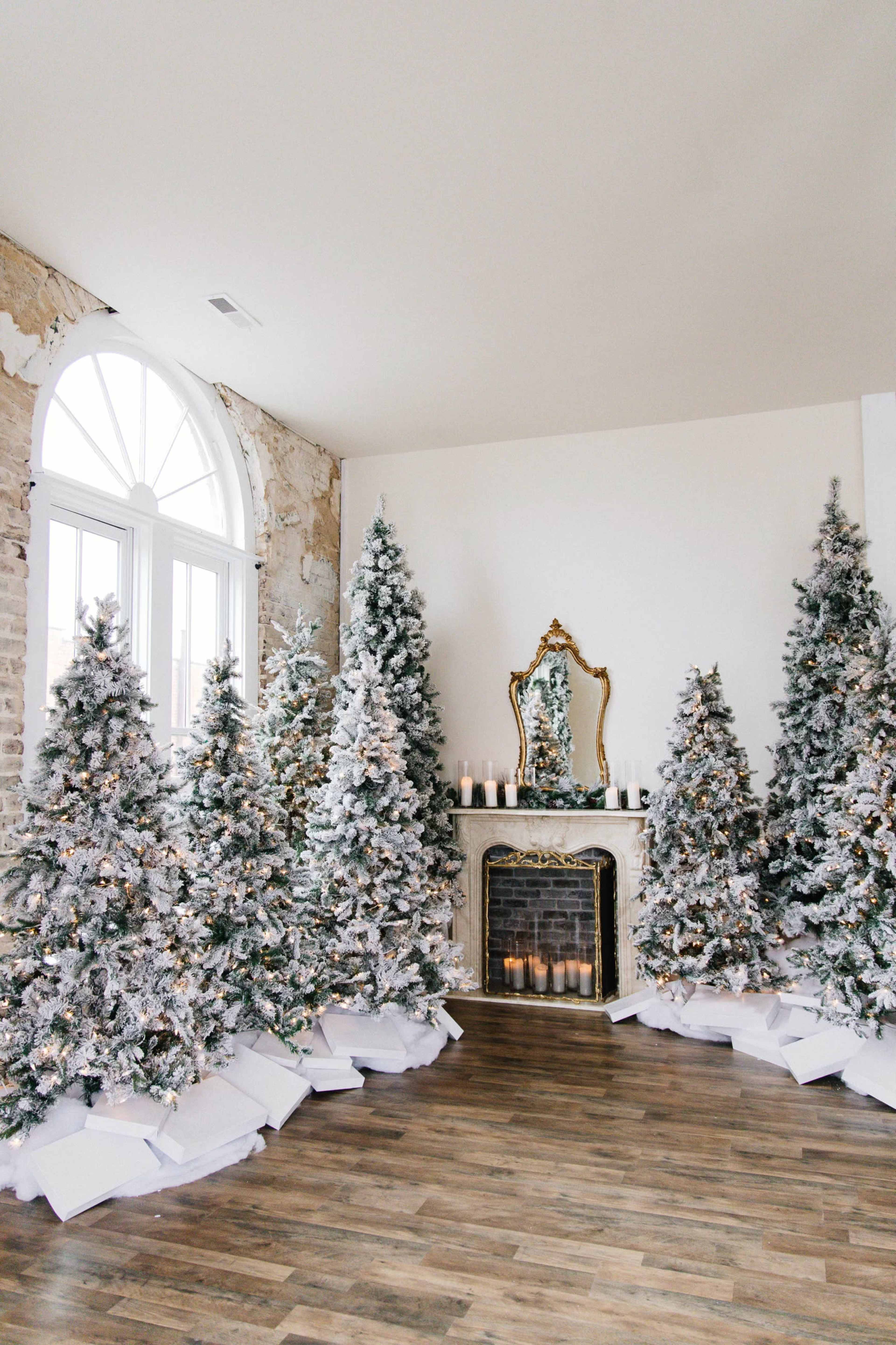 The scene features several decorated Christmas trees arranged around a fireplace, with a large mirror above it and wrapped gifts underneath the trees.