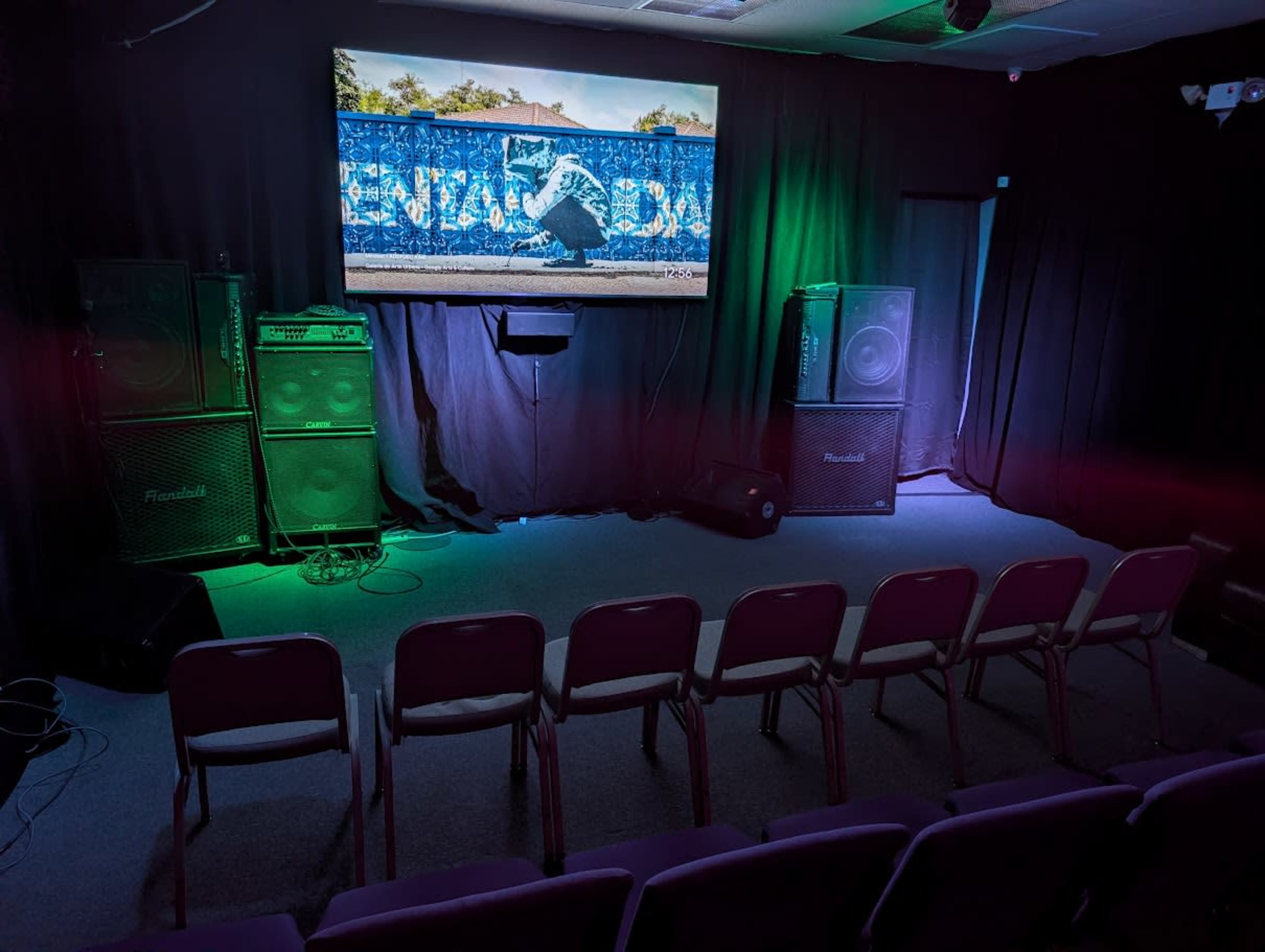 The image shows a small, dark room set up for a presentation, featuring multiple speakers, a large screen displaying an outdoor scene, and several rows of empty purple chairs.