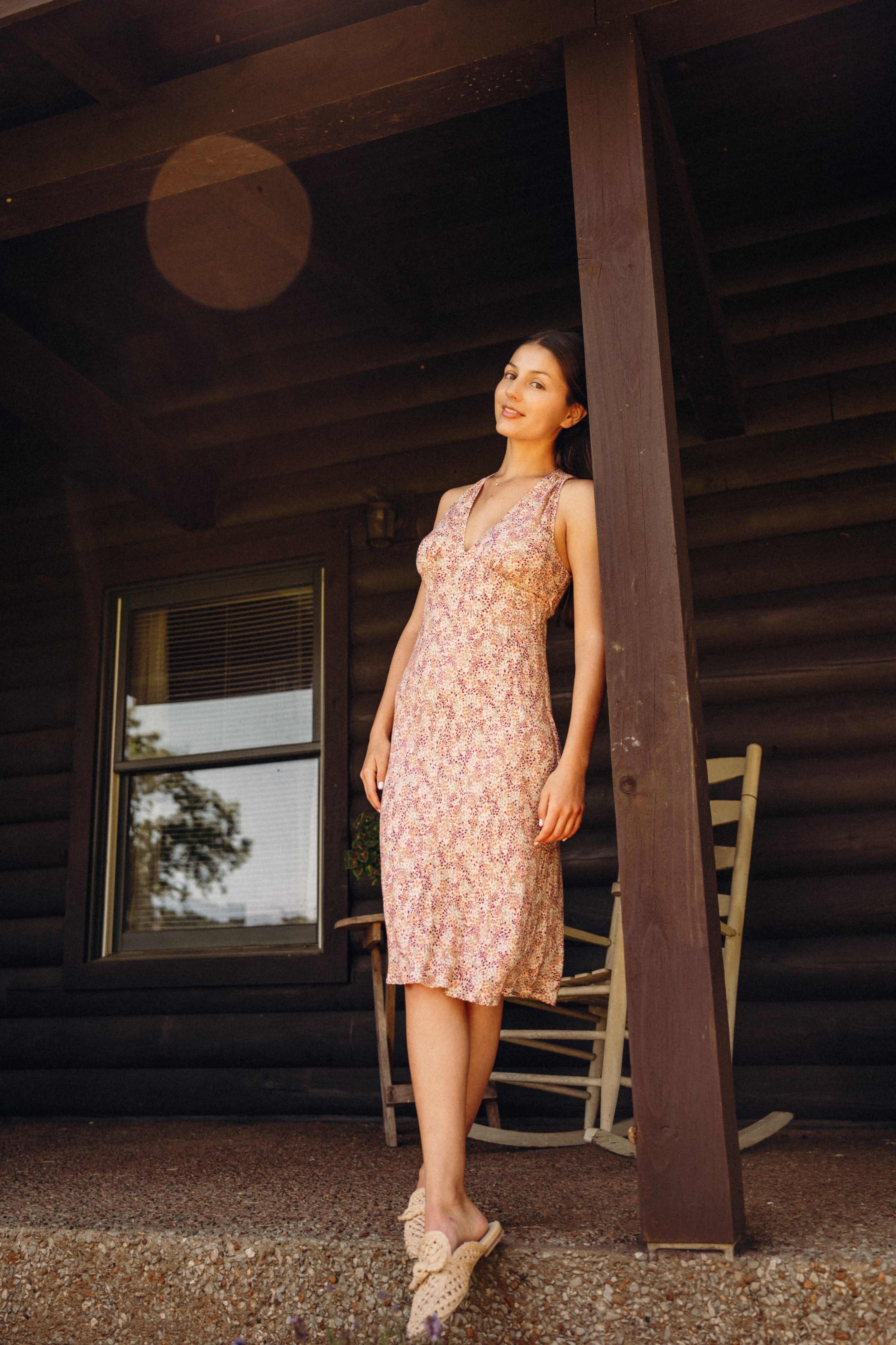 A woman stands on the porch of a log cabin, wearing a floral dress and facing the camera.
