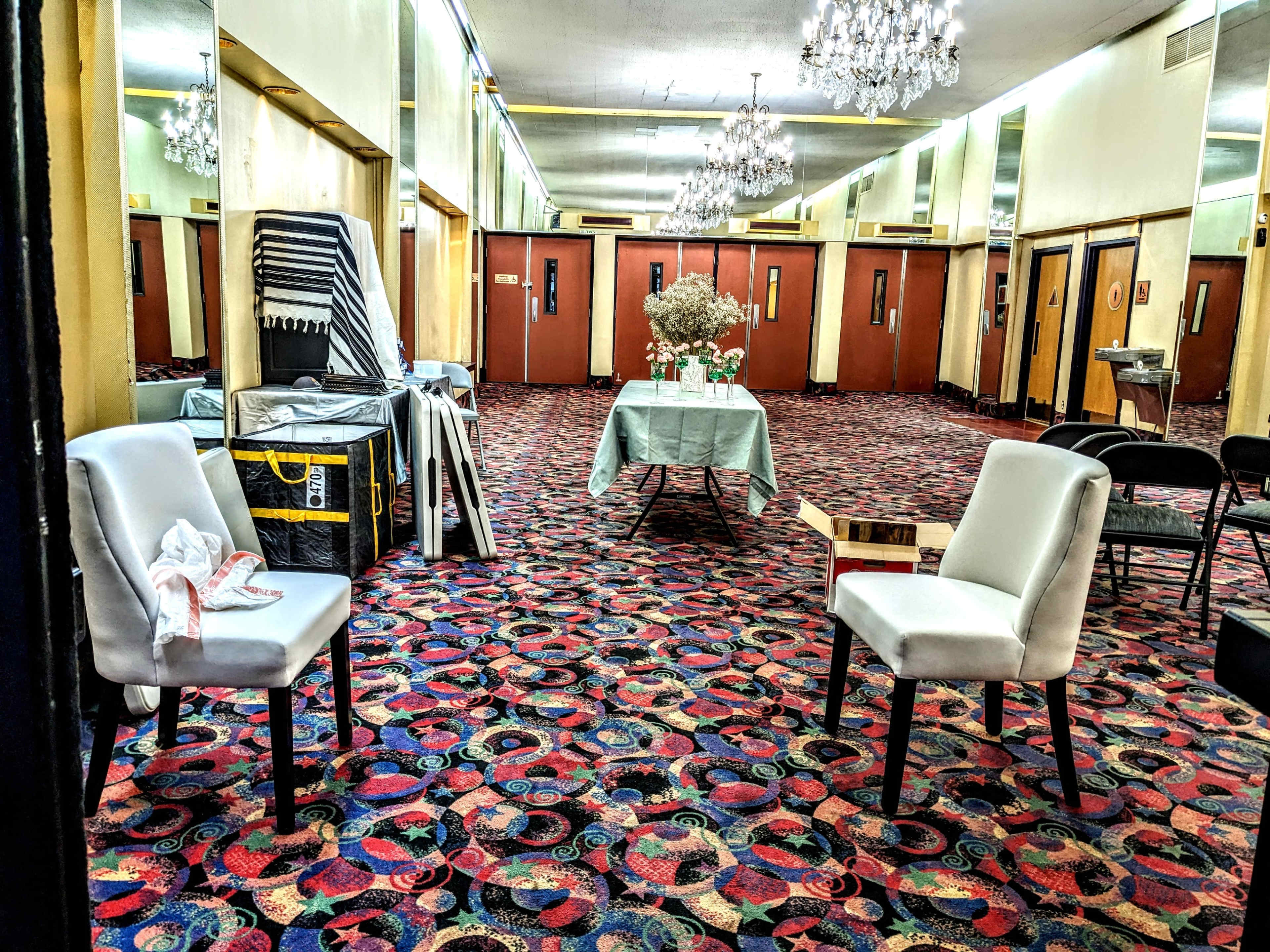 A brightly lit room with patterned carpeting, featuring two white chairs, a table set with a green tablecloth, and mirrors reflecting the surroundings, including a cart and several doors.