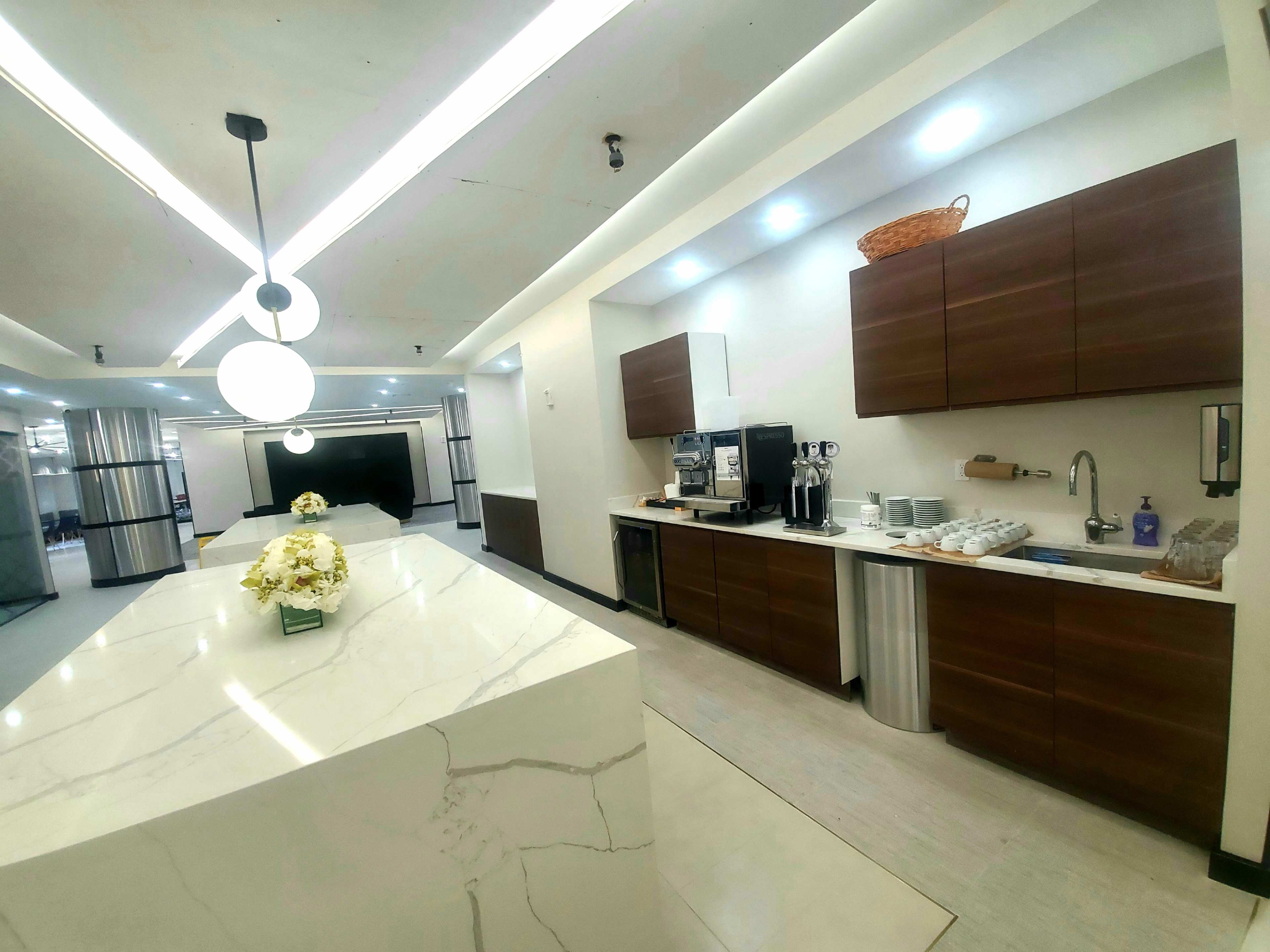 The image shows a modern kitchen area featuring a marble countertop, wooden cabinetry, and coffee machines along with a flower arrangement on the counter.