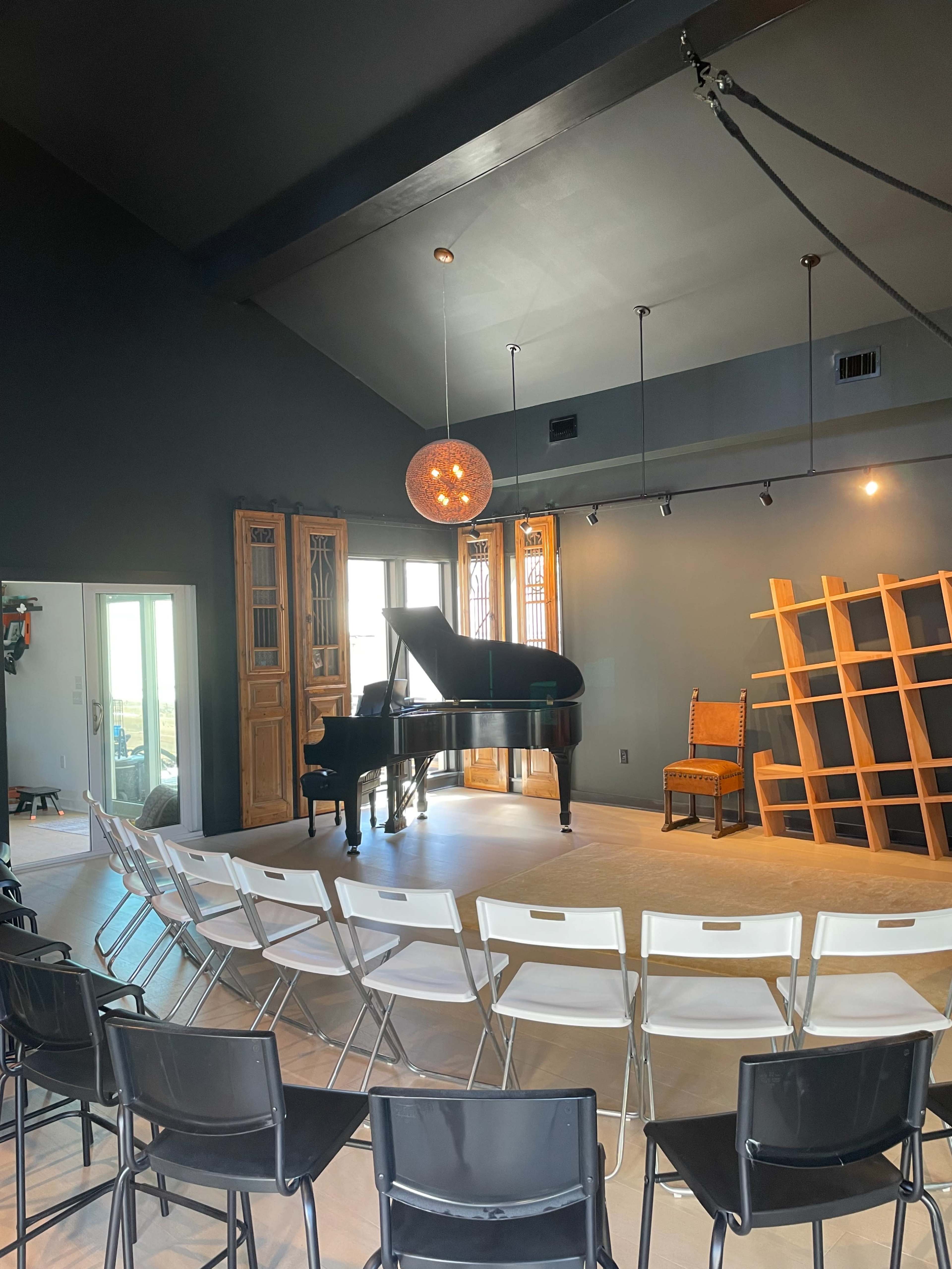 A grand piano is positioned on a wooden stage, surrounded by rows of black and white chairs in a room with dark walls and large windows.