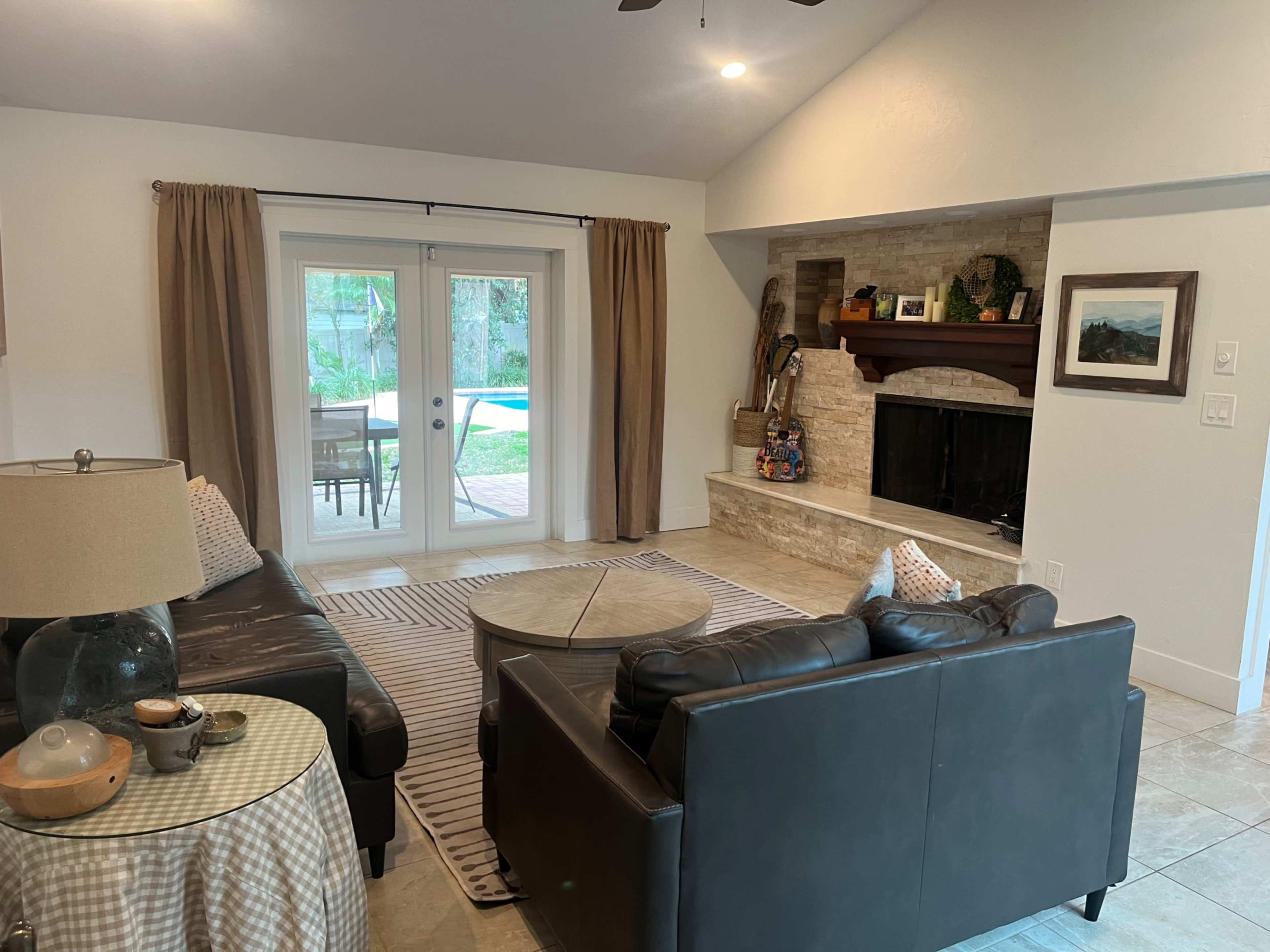 A cozy living room featuring two black sofas facing a coffee table, with a stone fireplace and glass doors leading to a patio area with a pool.
