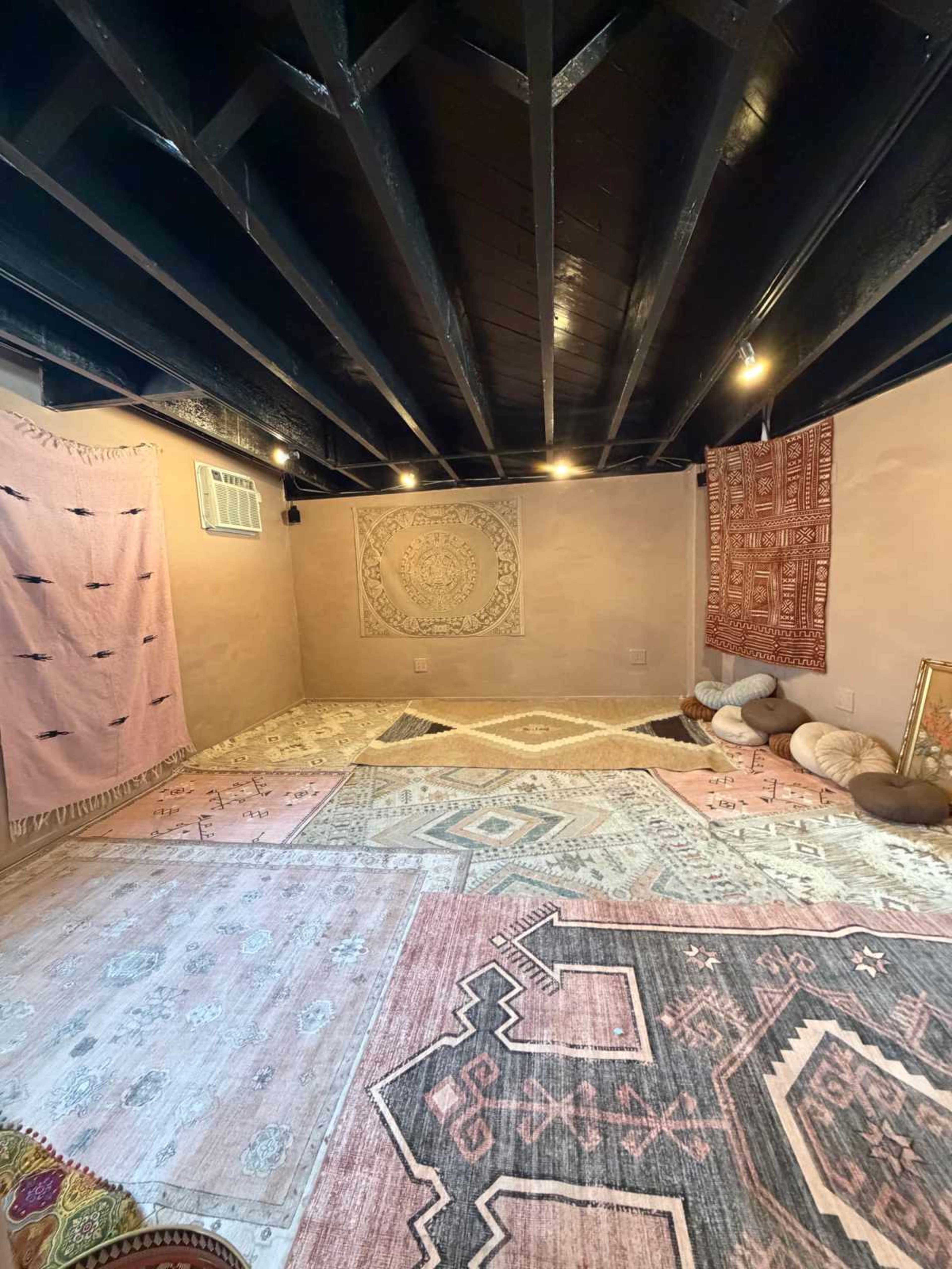 The image shows a spacious, softly lit room with a variety of patterned rugs on the floor, decorated walls, and several floor cushions.