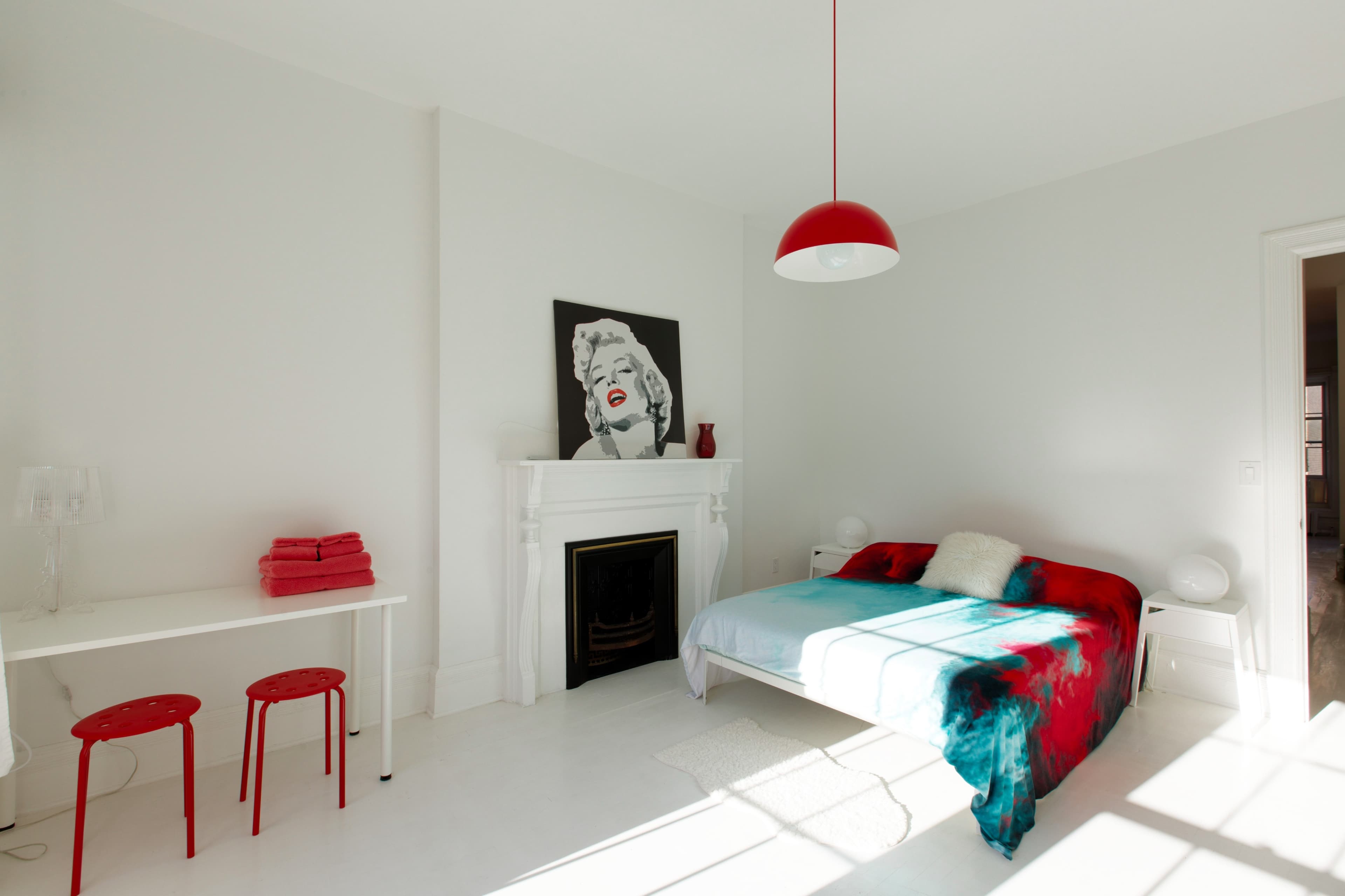 The bedroom features a white color scheme with a red accent lamp, a bed with a colorful throw, a small table with red stools, and a large black-and-white artwork on the wall.
