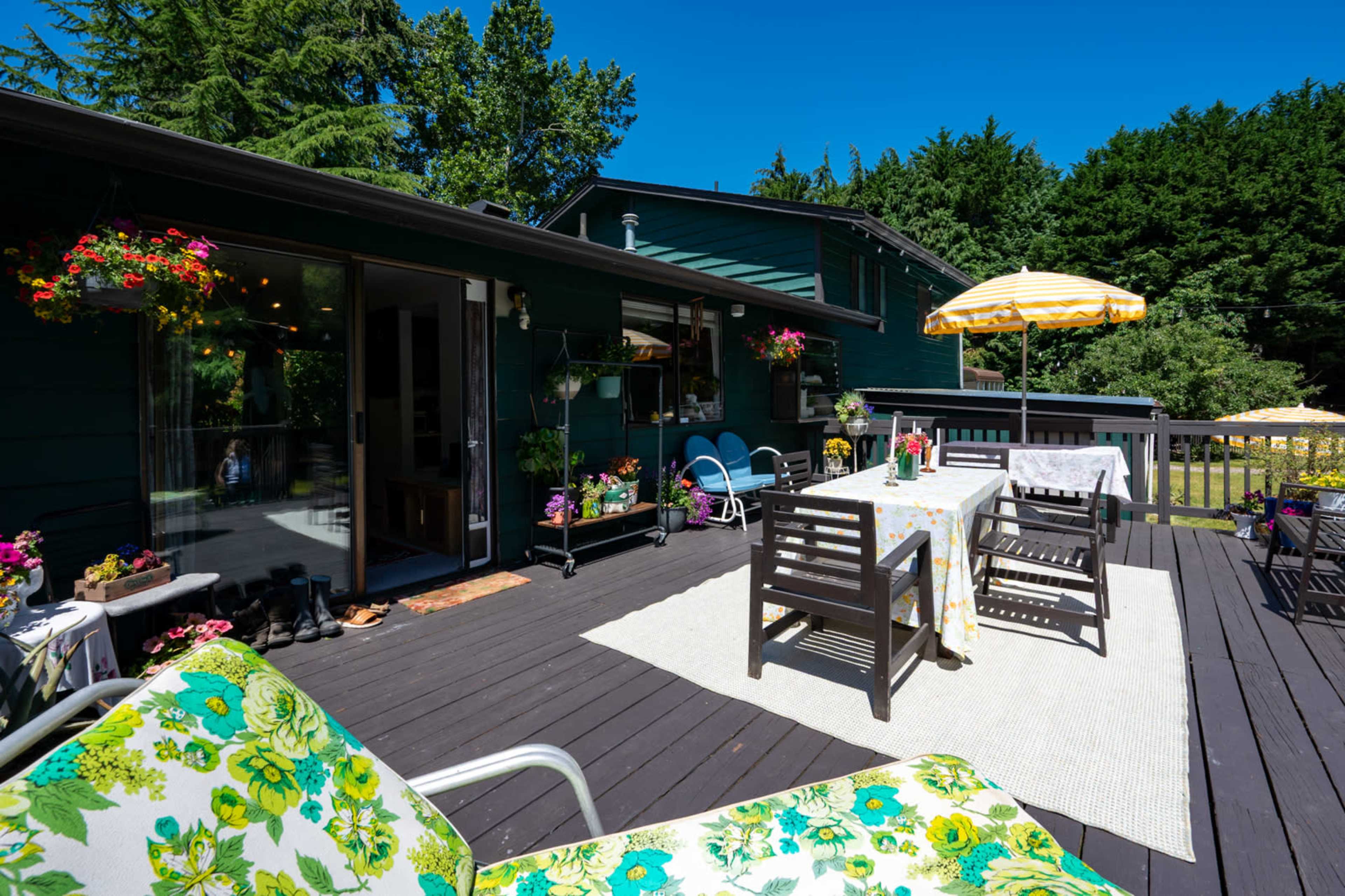A spacious wooden deck features outdoor seating, dining tables, and colorful floral decorations, with a house and lush greenery in the background.