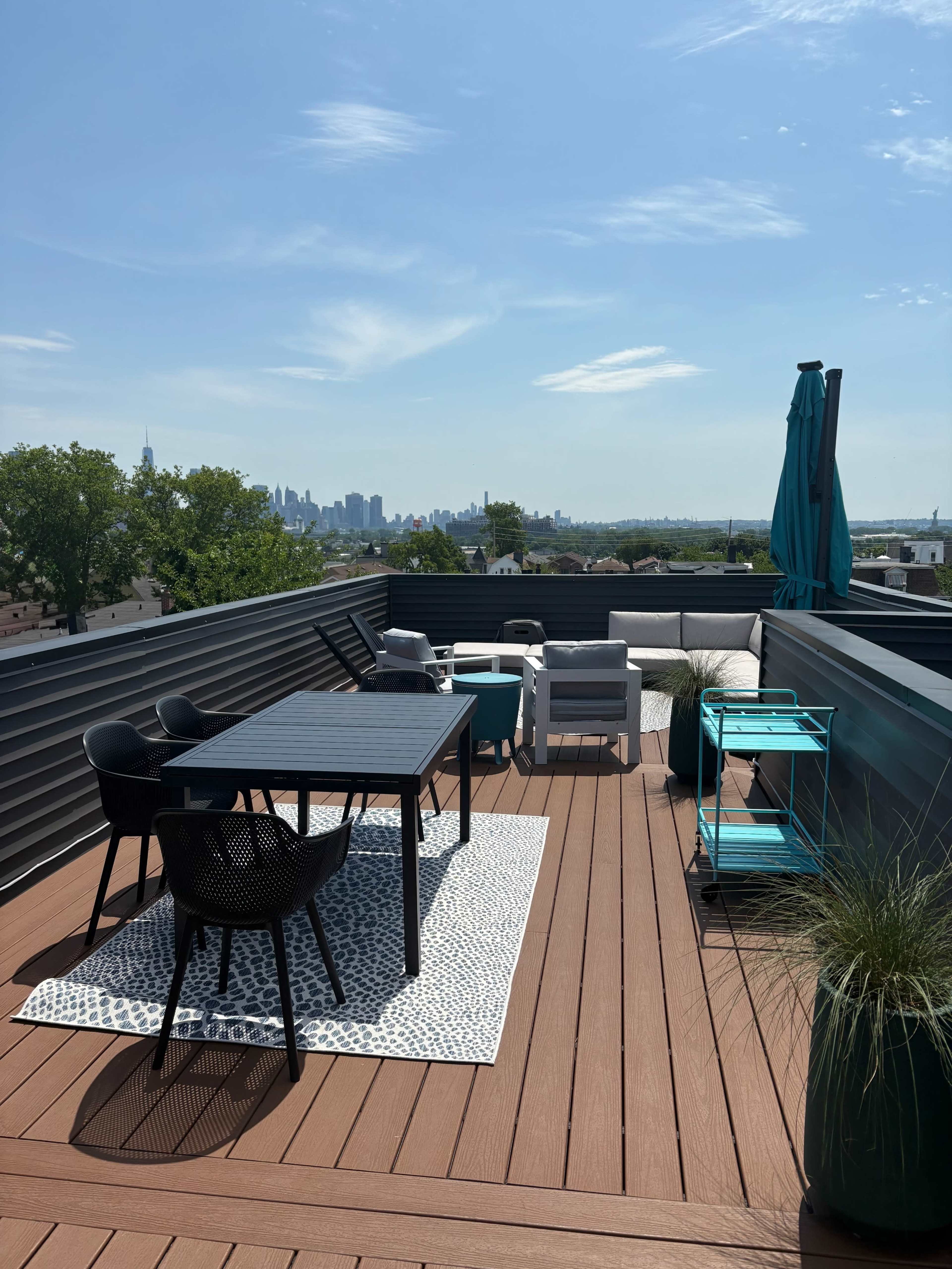 The image shows a rooftop terrace featuring a black table, several chairs, and a lounge area, with a view of the city skyline in the background.
