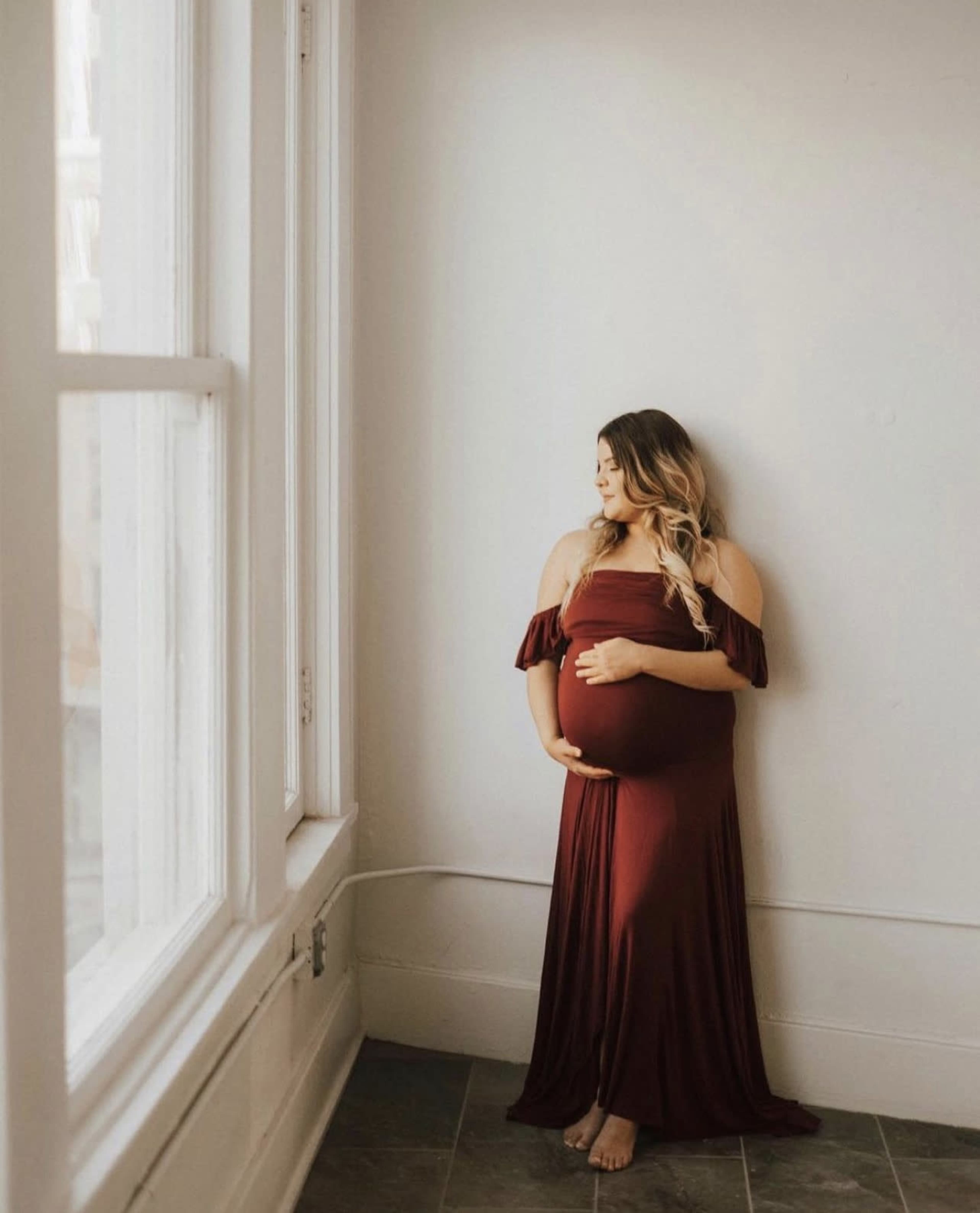 A pregnant woman in a maroon off-the-shoulder dress stands by a large window against a plain wall, holding her belly.