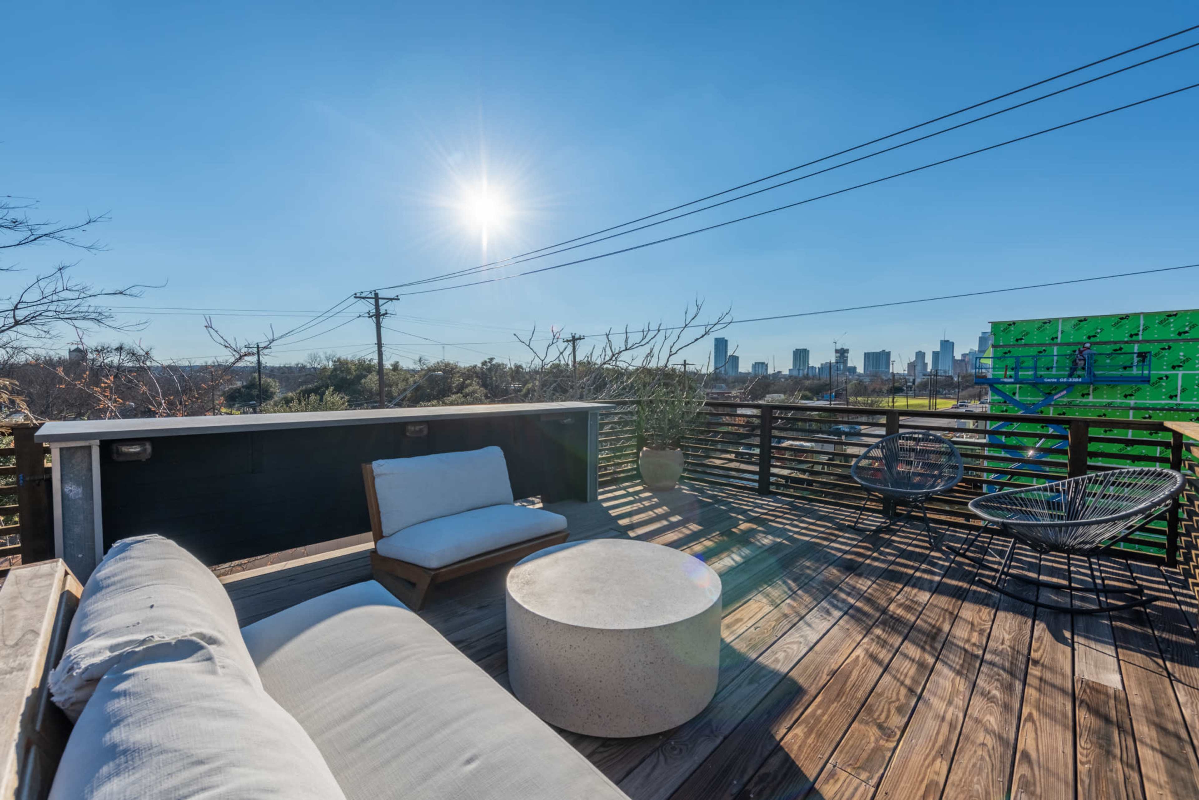 A rooftop terrace features a seating area with a concrete table and views of a city skyline under a clear blue sky.