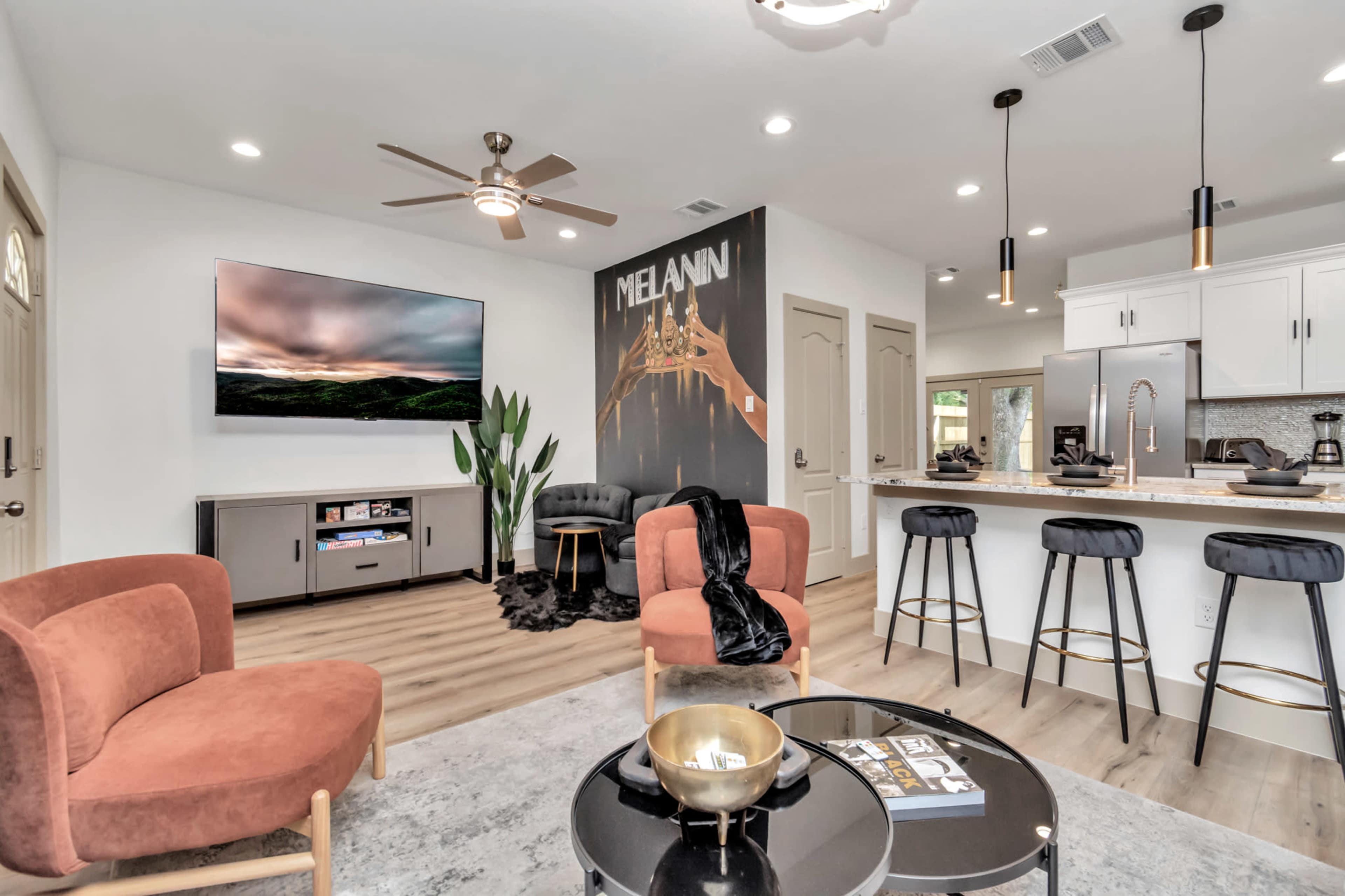 A modern living room features an open layout with a television on the wall, stylish furniture, and a kitchen area in the background.