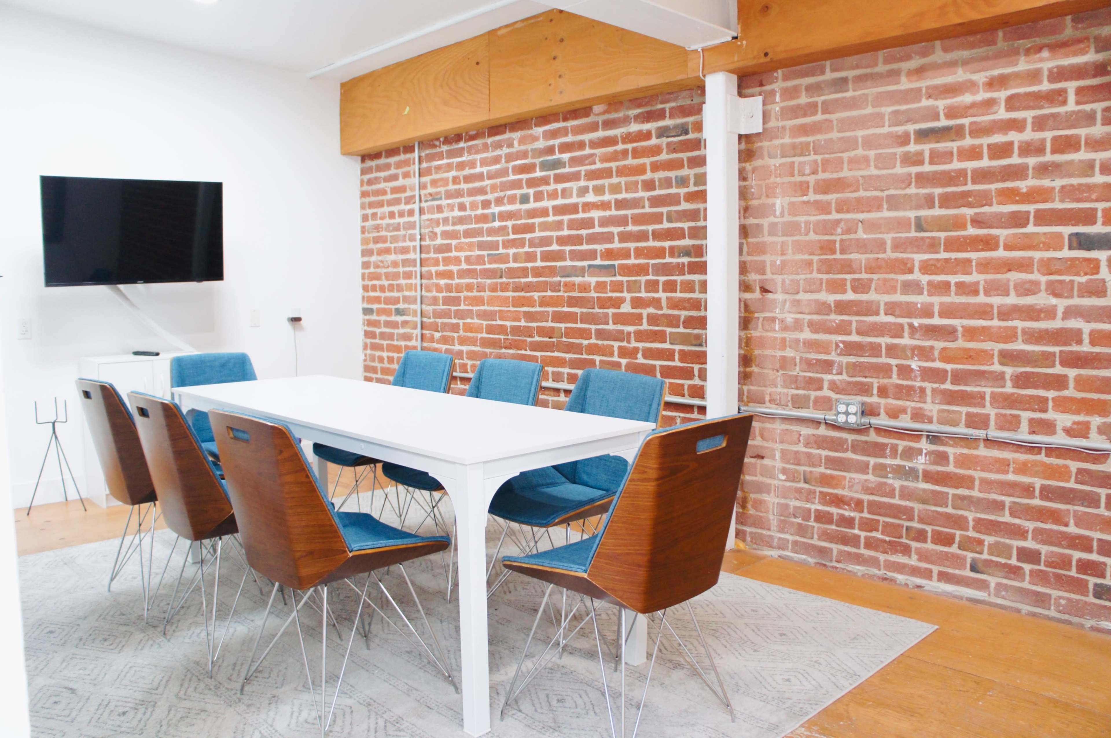 A rectangular white table surrounded by six blue chairs is set against a backdrop of exposed brick walls in a well-lit room.