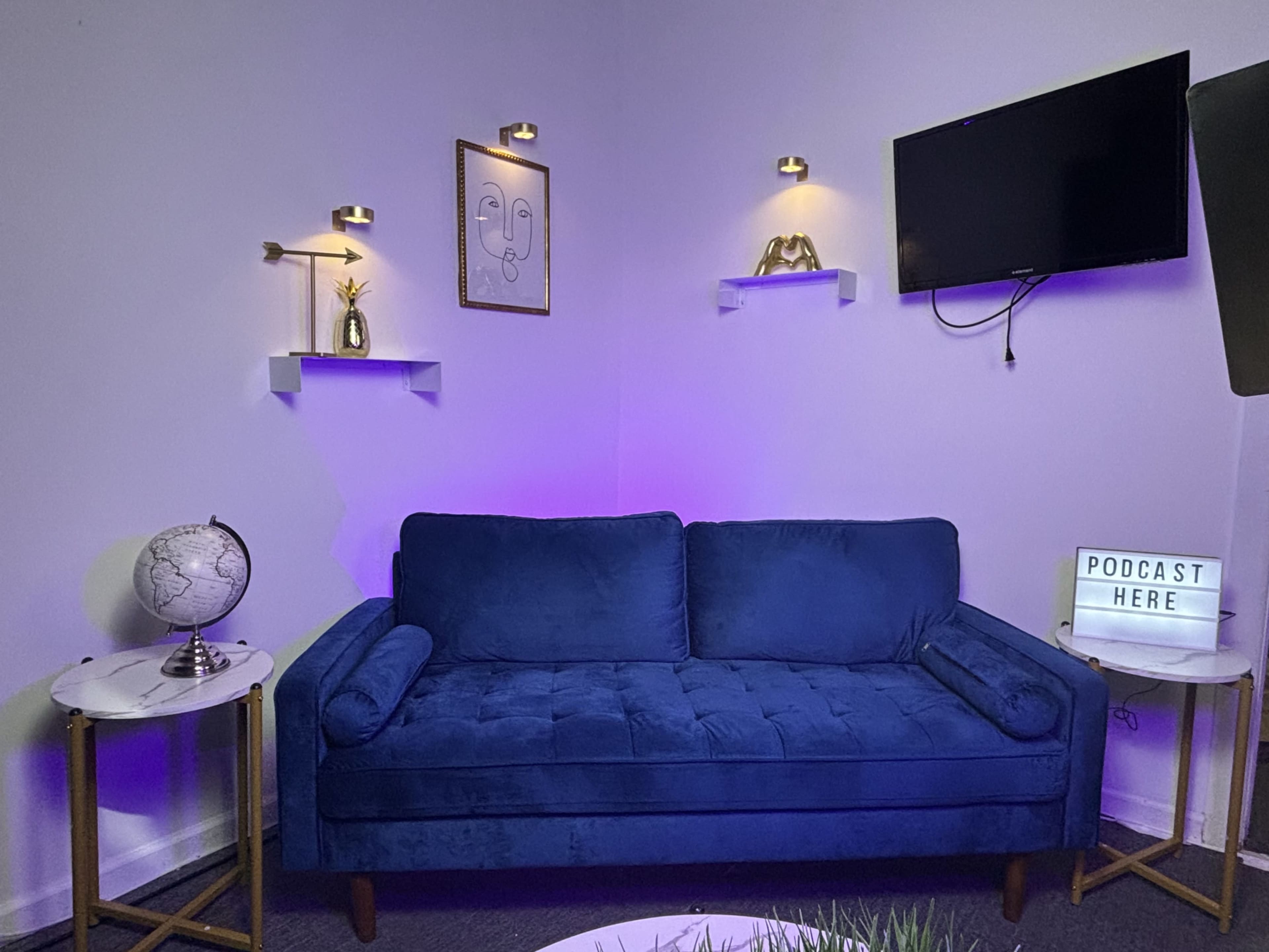 The image shows a cozy podcast studio with a blue velvet sofa, two side tables, a globe, decorative wall art, and modern lighting.