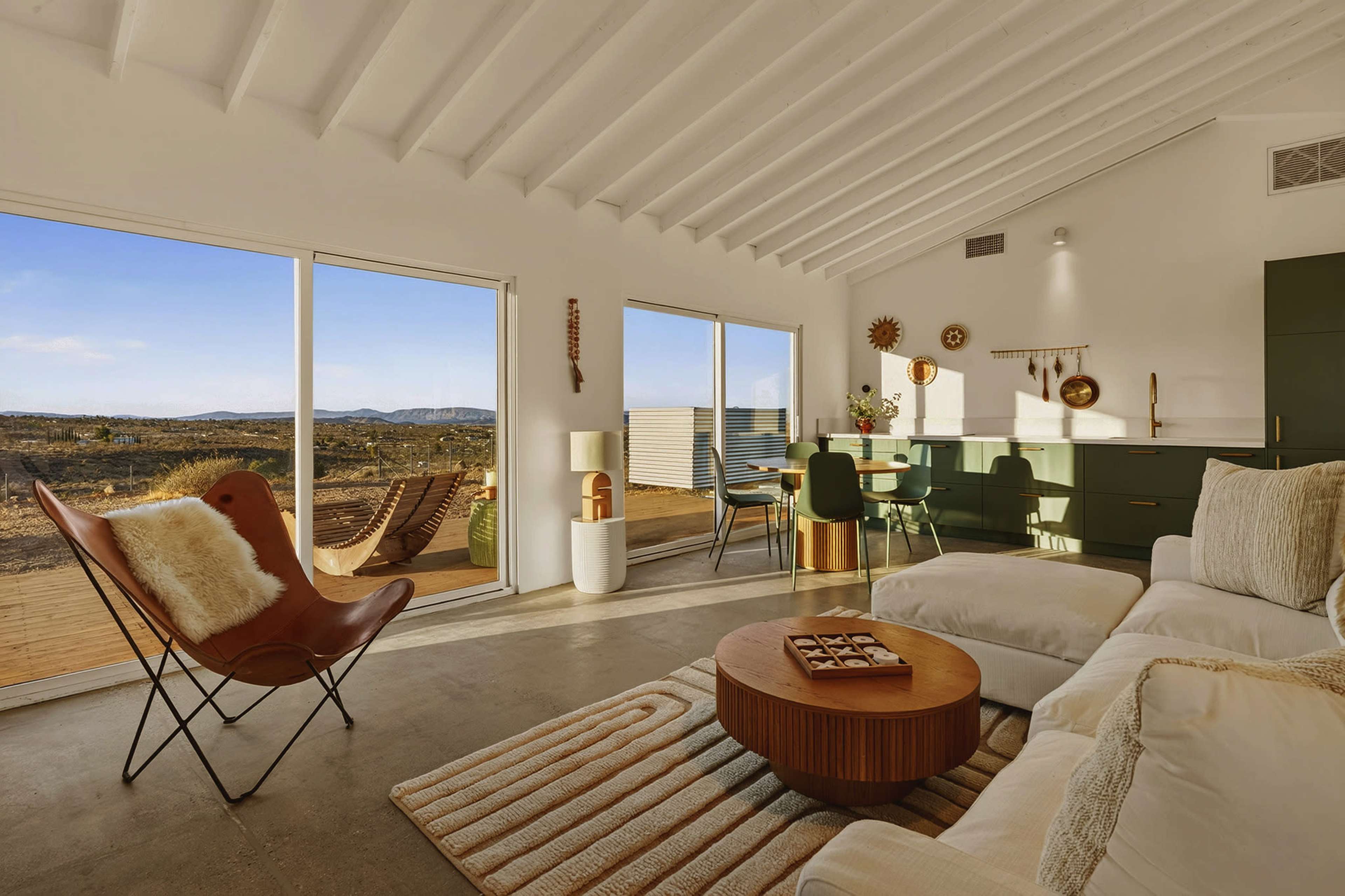 The image shows a modern living room with large windows overlooking a desert landscape, featuring a white sofa, a round coffee table, and a dining area with green chairs.