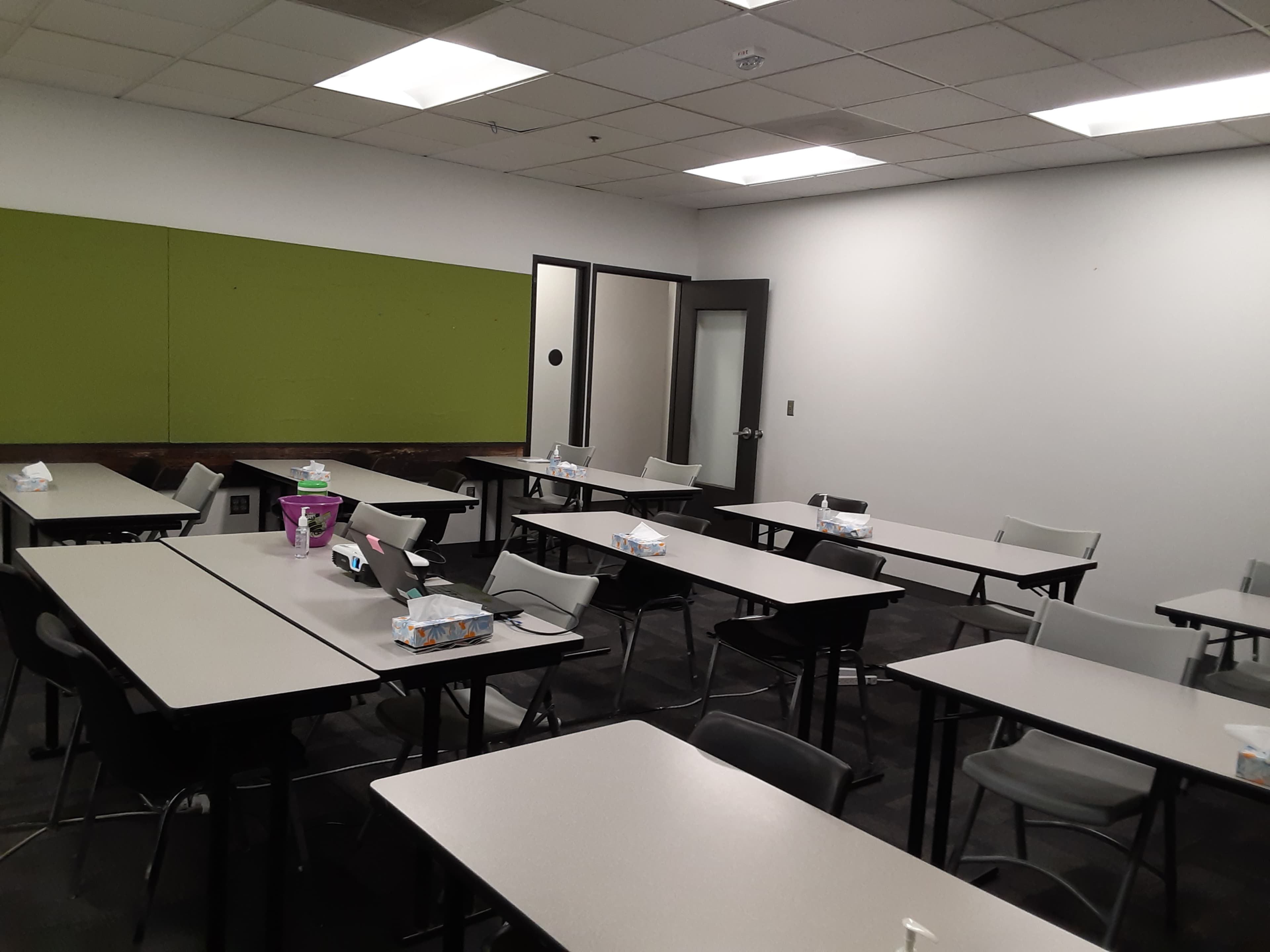 The image shows a classroom with several tables arranged in a grid pattern, each equipped with chairs and a few items on the tables.