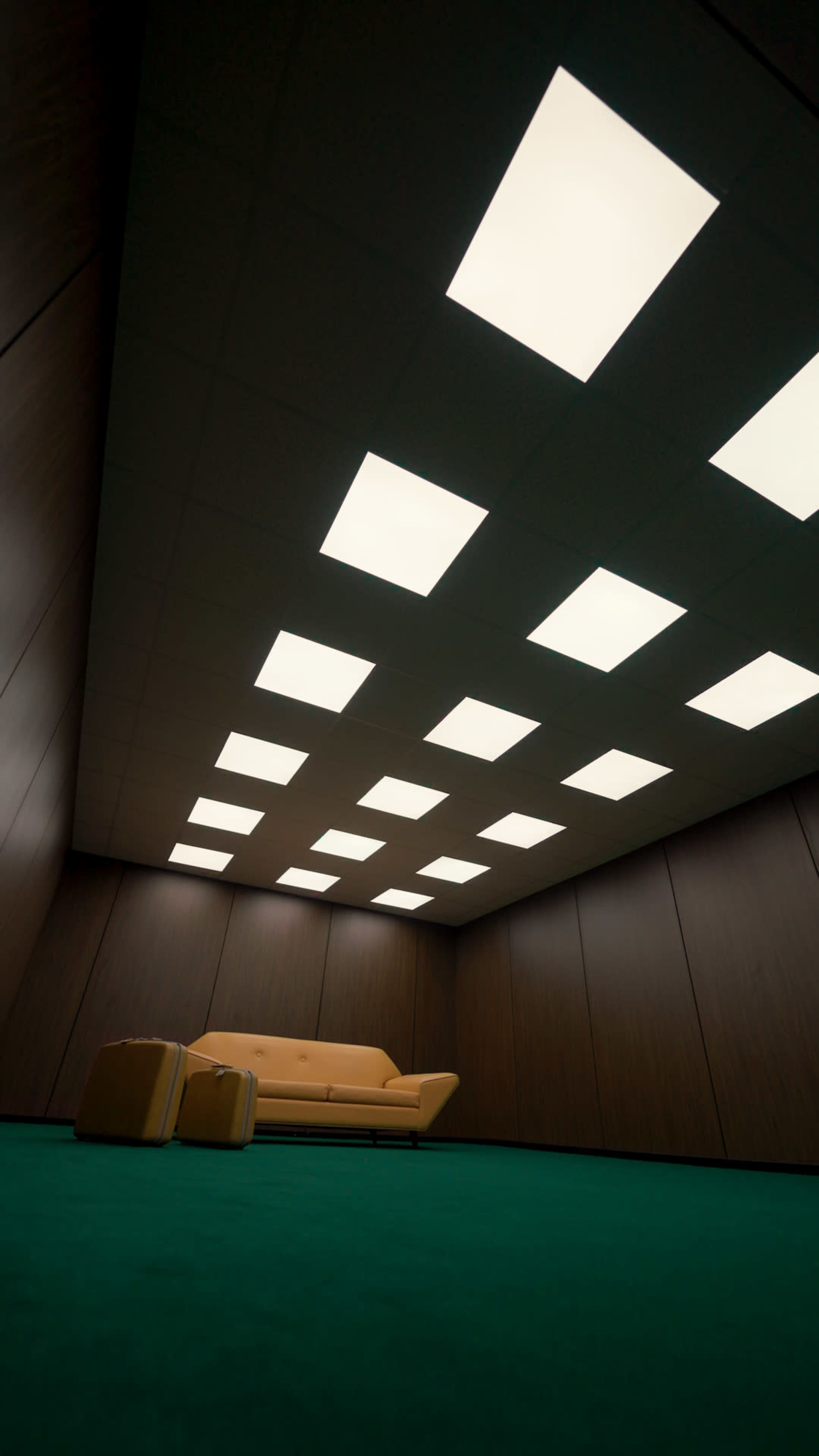The image shows a sparsely furnished room with a yellow sofa and a green carpet, illuminated by rectangular ceiling lights.