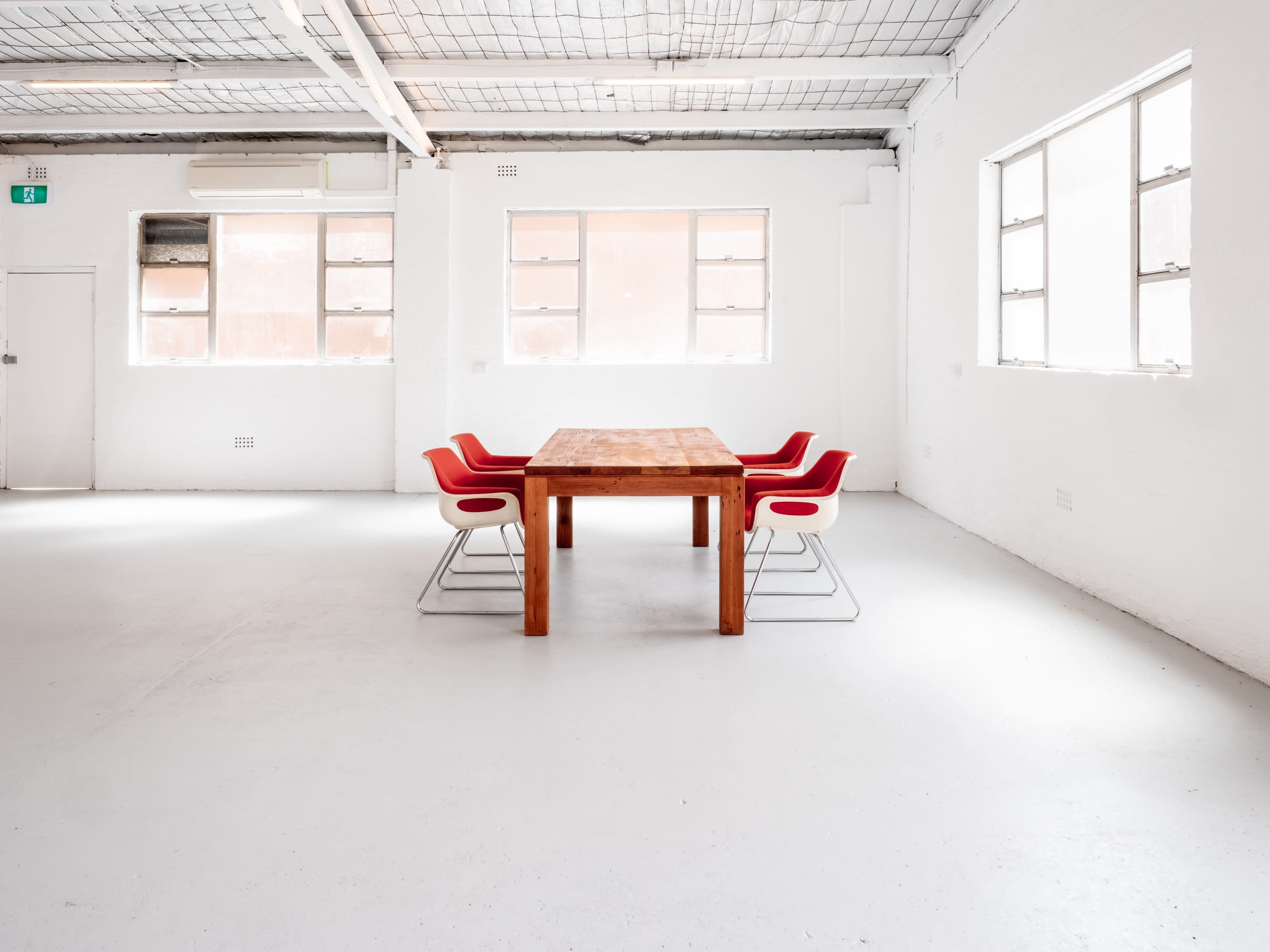 A minimalist meeting room features a wooden table surrounded by four red chairs, with large windows letting in natural light.