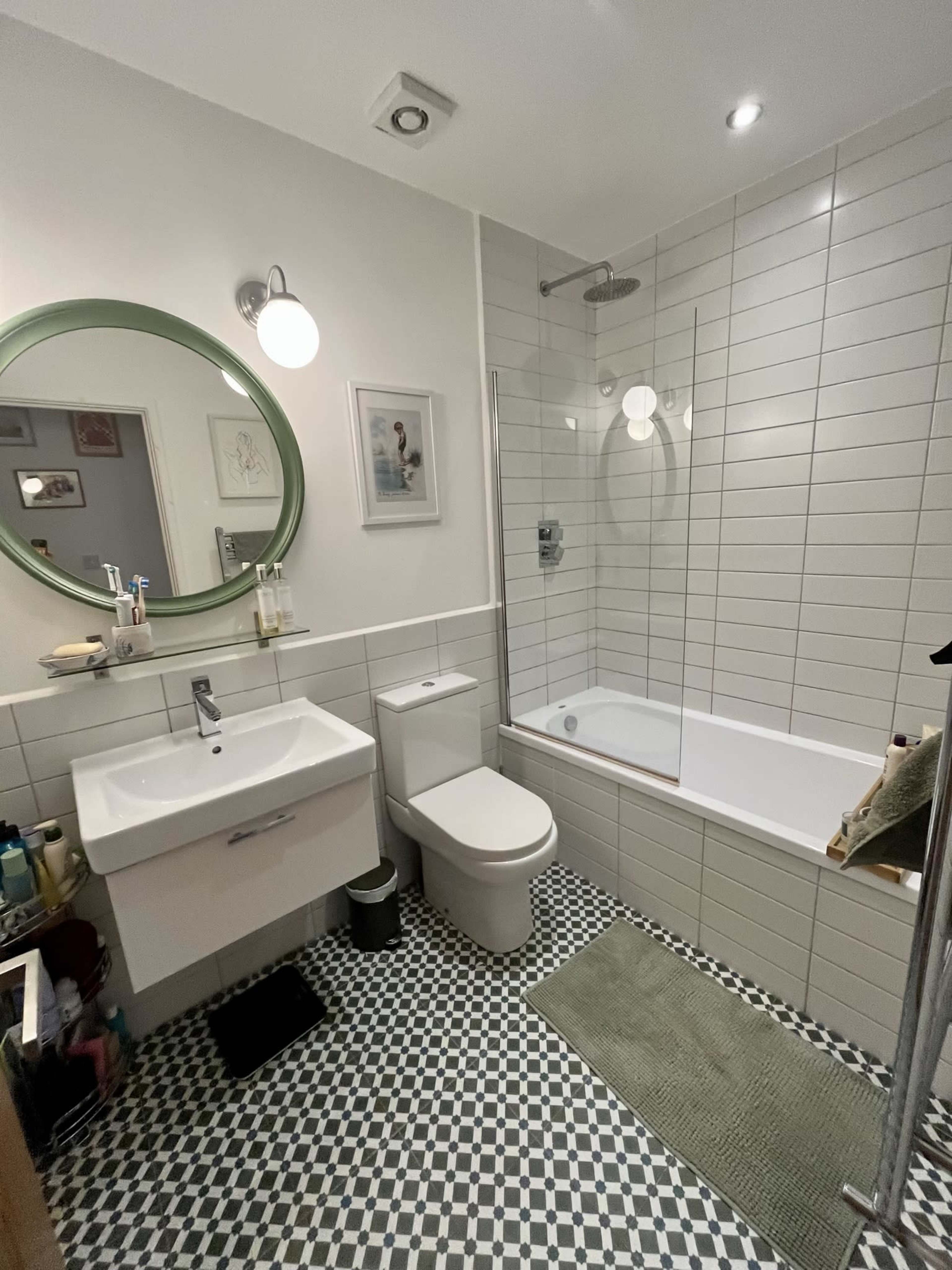 A modern bathroom featuring a shower-over-bath setup, a wall-mounted sink with storage underneath, a toilet, and a circular mirror above the sink, all arranged with patterned tile flooring.