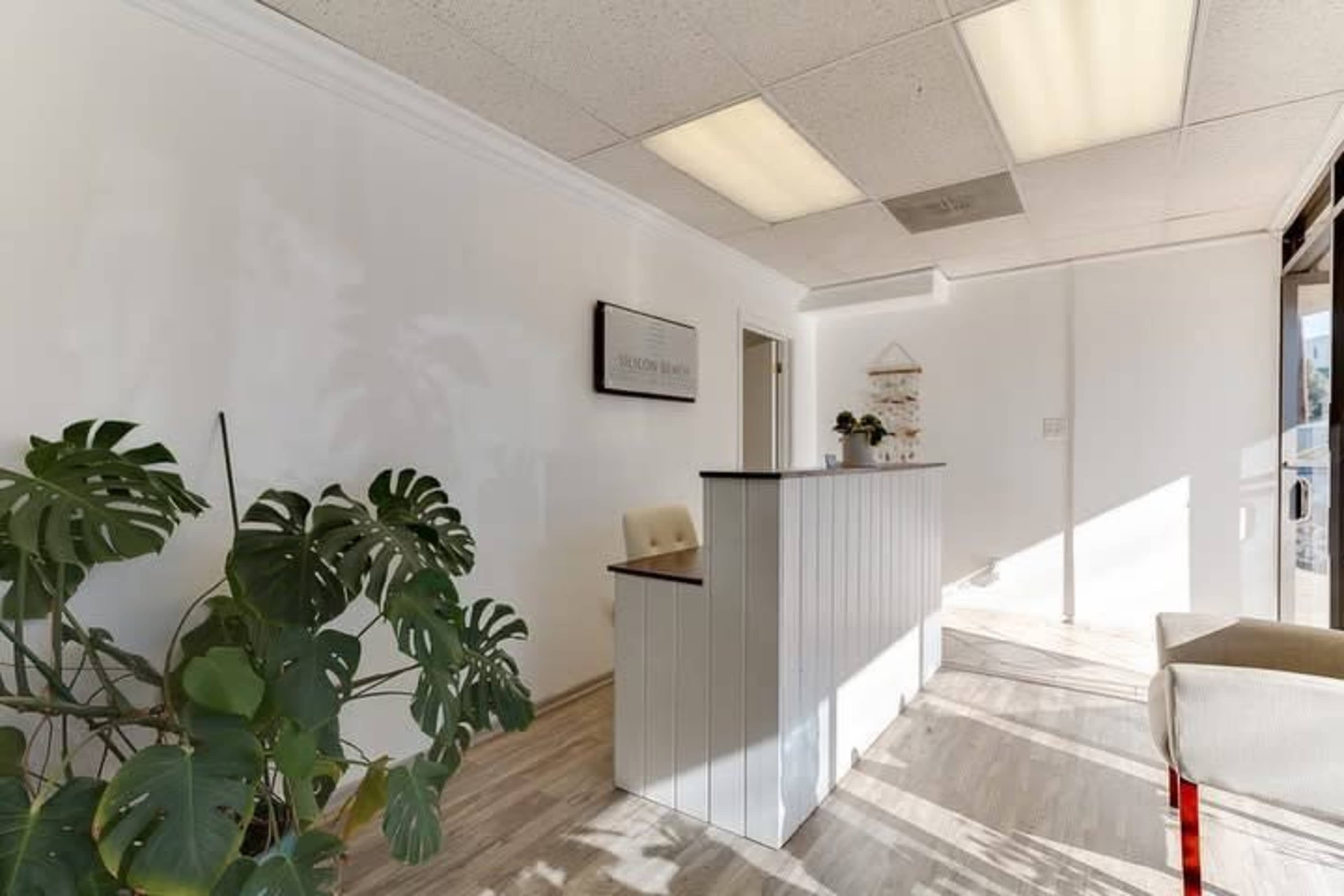 The image shows a bright, minimalist reception area with a white desk and a large plant in the corner.