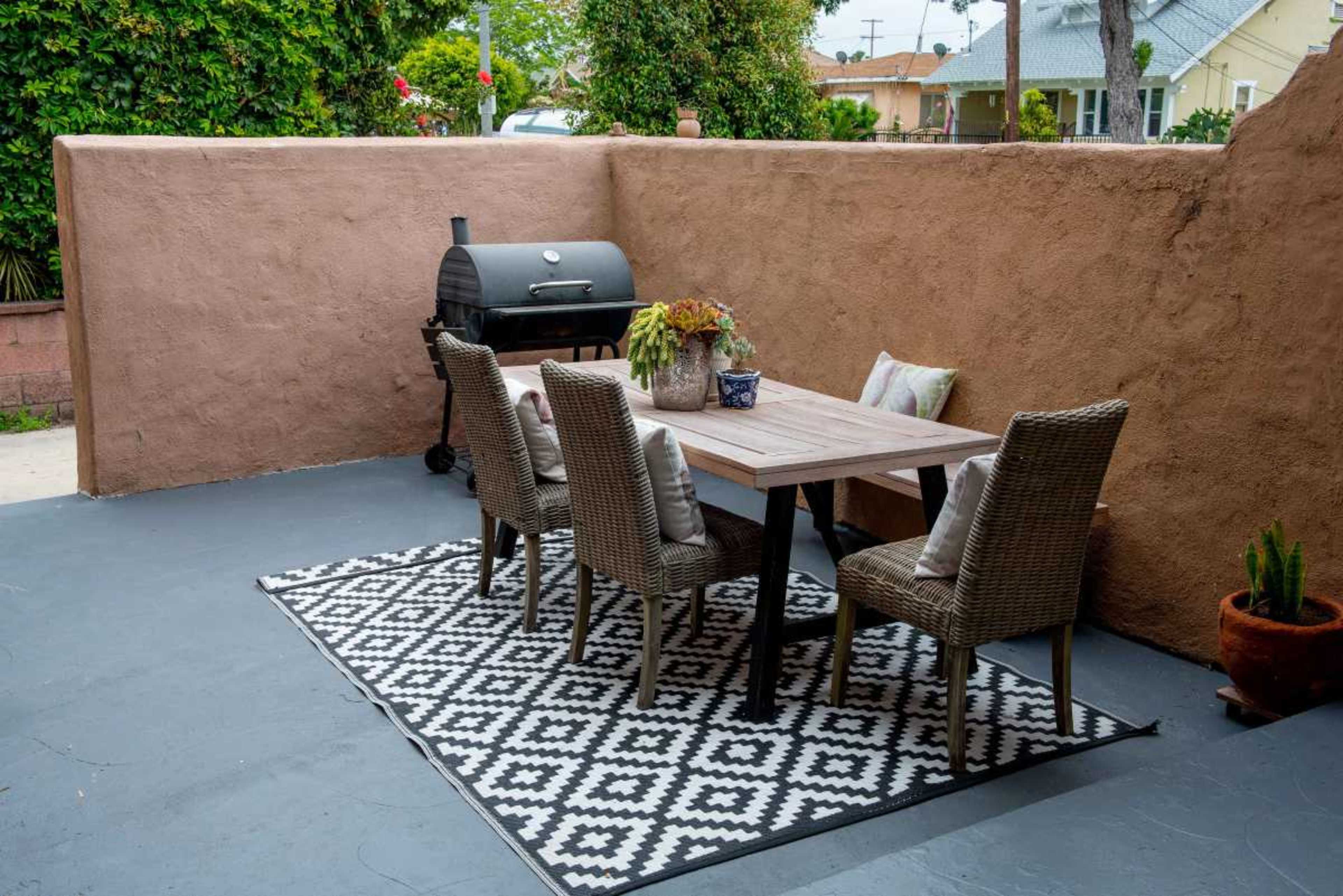 The image shows a patio area featuring a wooden dining table surrounded by four wicker chairs, a patterned rug underneath, and a barbecue grill against a textured wall.