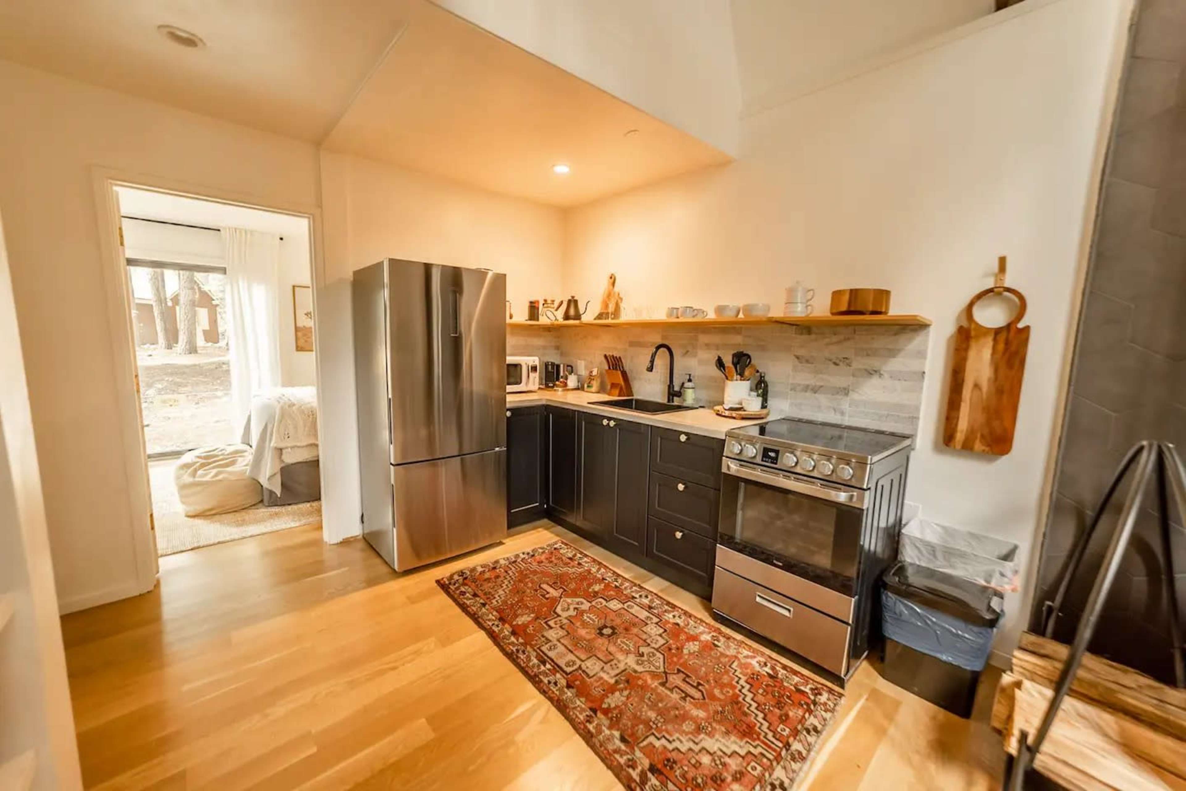 A modern kitchen features stainless steel appliances, dark cabinetry, and a decorative rug on a hardwood floor, with a doorway leading to a living area.