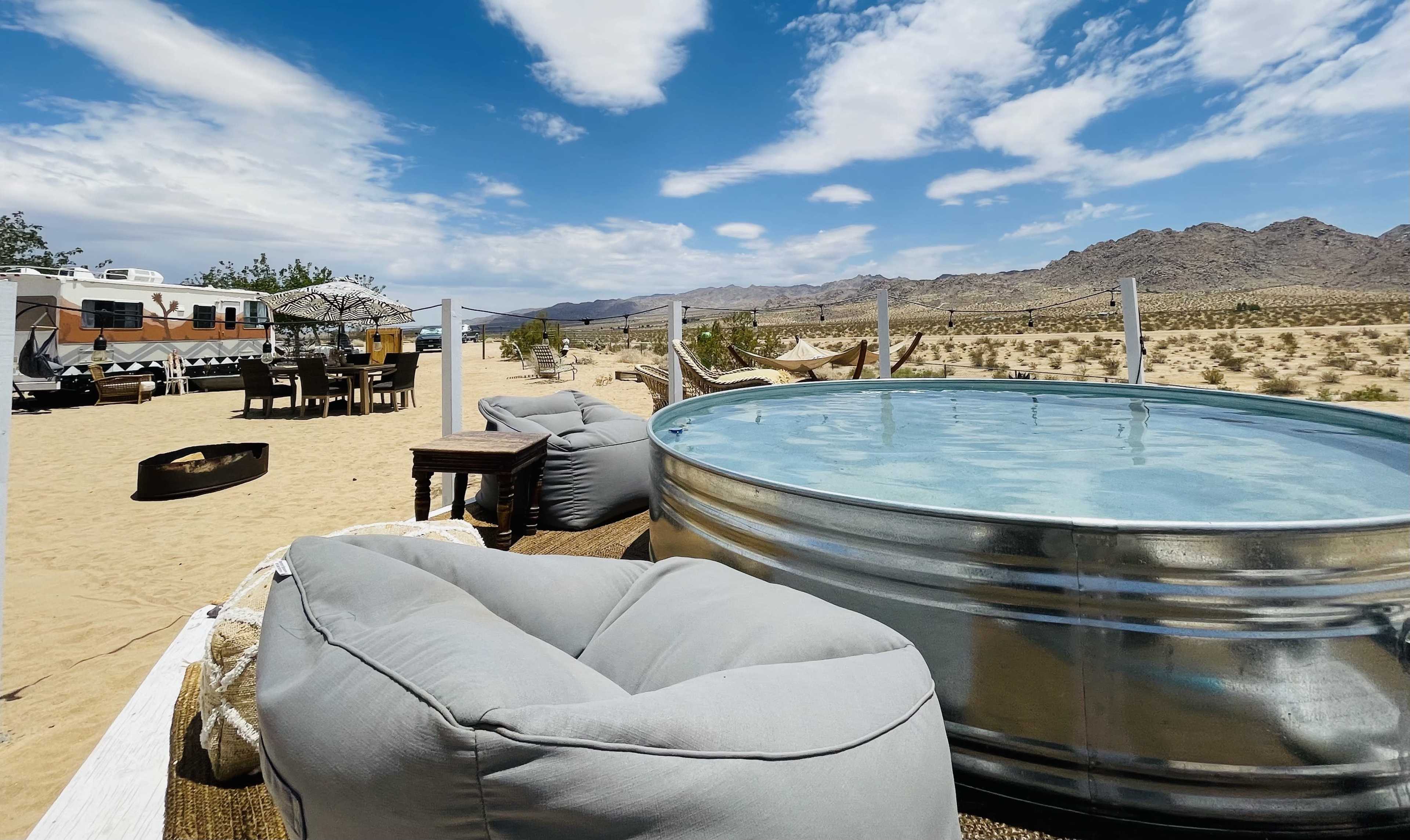 A metal pool sits beside a seating area with bean bags and a dining set against a desert backdrop with mountains in the distance.