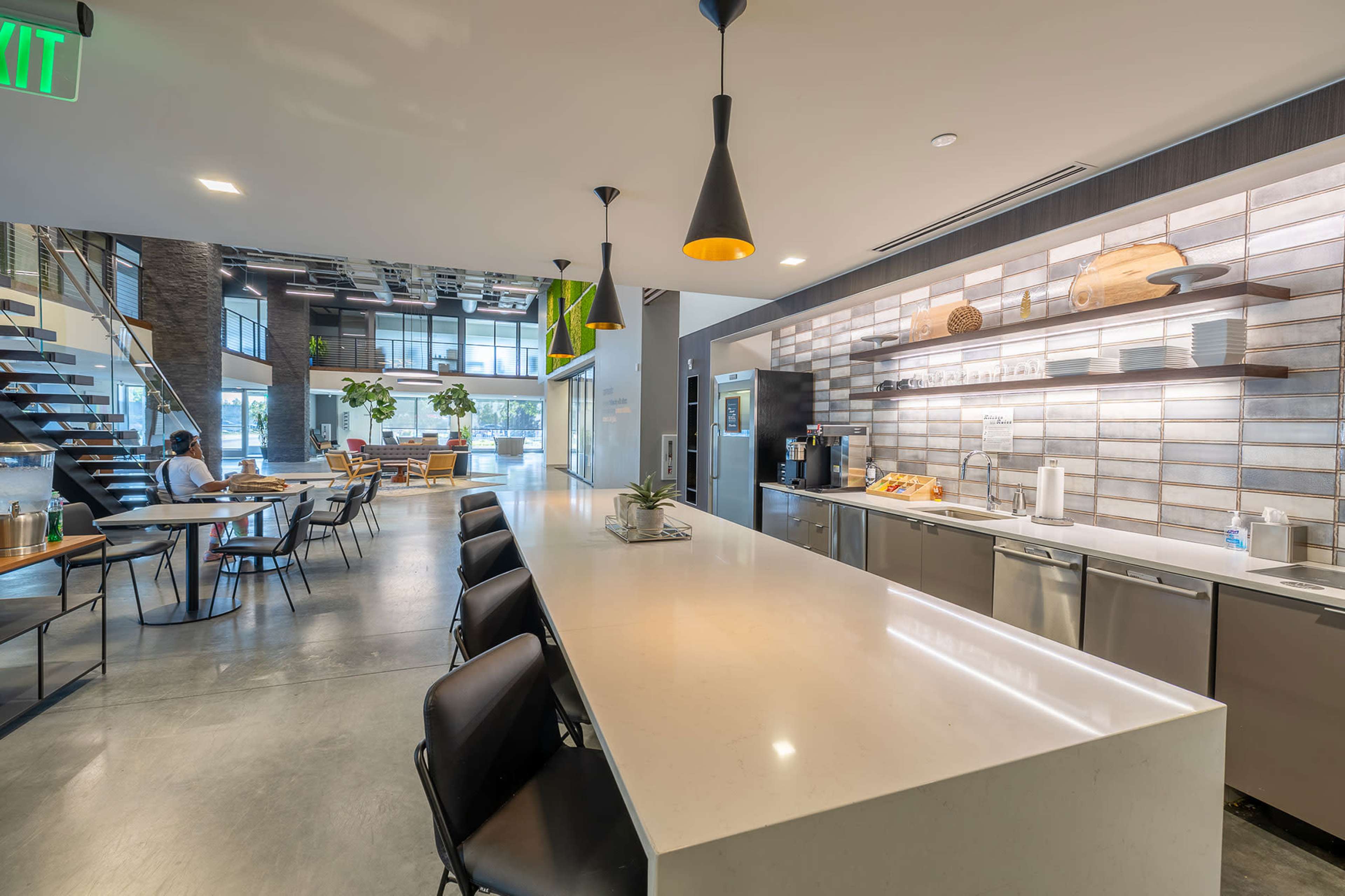 The image shows a modern kitchen area with a long white countertop, black chairs, and a casual dining space in the background featuring plants and seating.