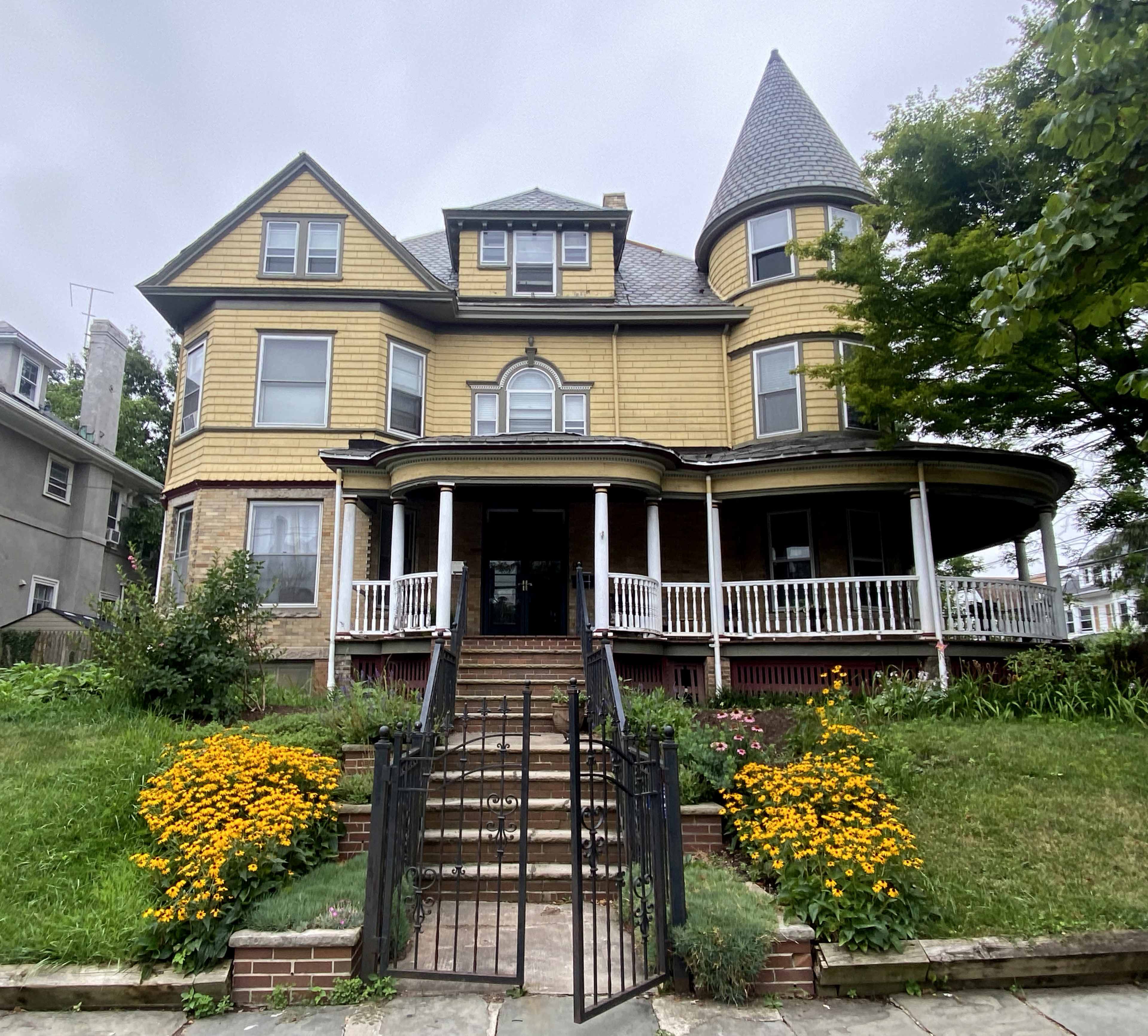 A large, multi-story Victorian house with a conical tower and a wraparound porch is surrounded by a garden featuring bright yellow flowers.