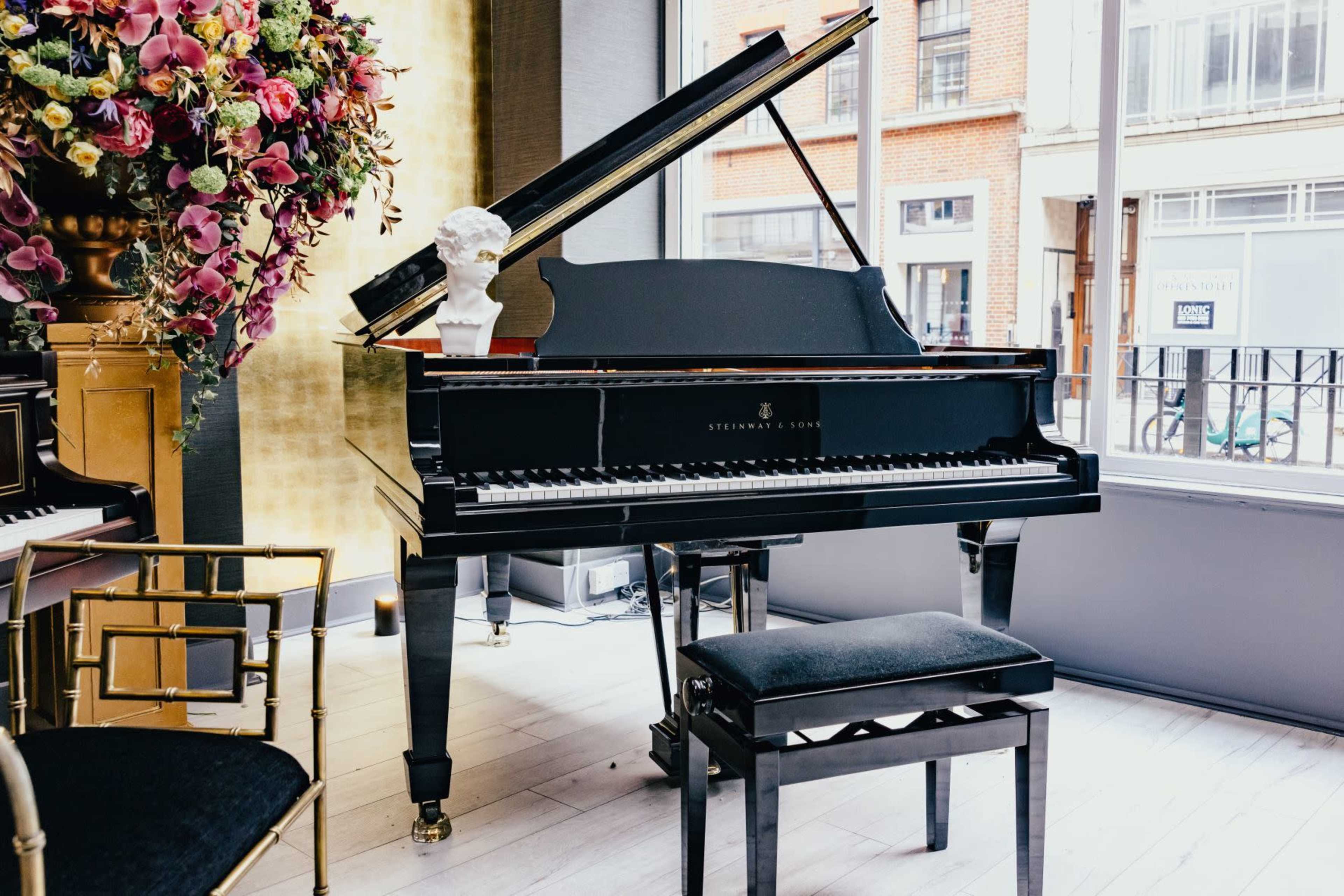 A black grand piano sits beside a large window, adorned with a floral arrangement and a decorative bust.