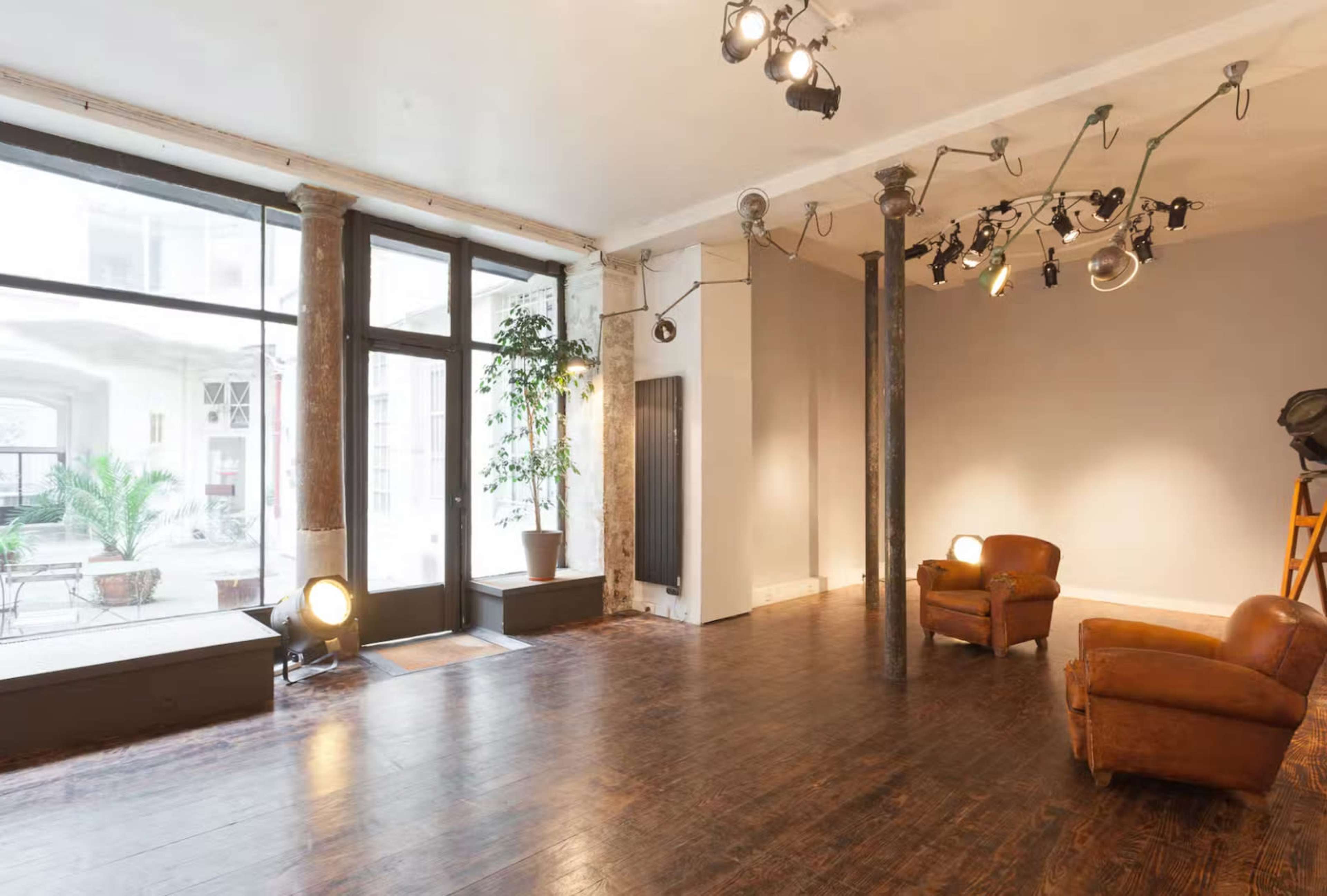 The image shows a spacious room with wooden flooring, two brown leather chairs, large windows, and a lighting setup on the ceiling.