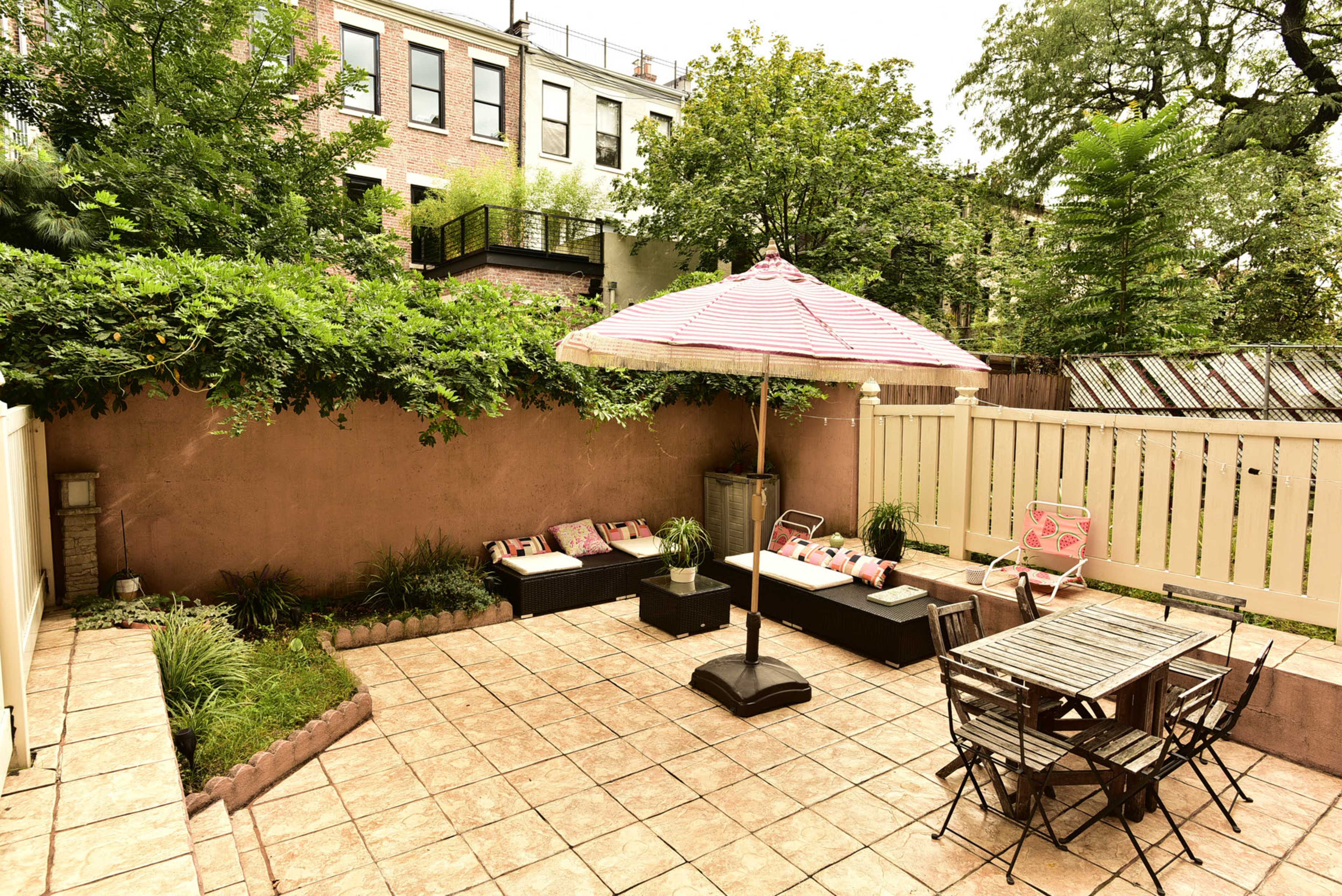 The image depicts a fenced outdoor patio area featuring a dining table, lounge seating, and a large umbrella, surrounded by greenery.