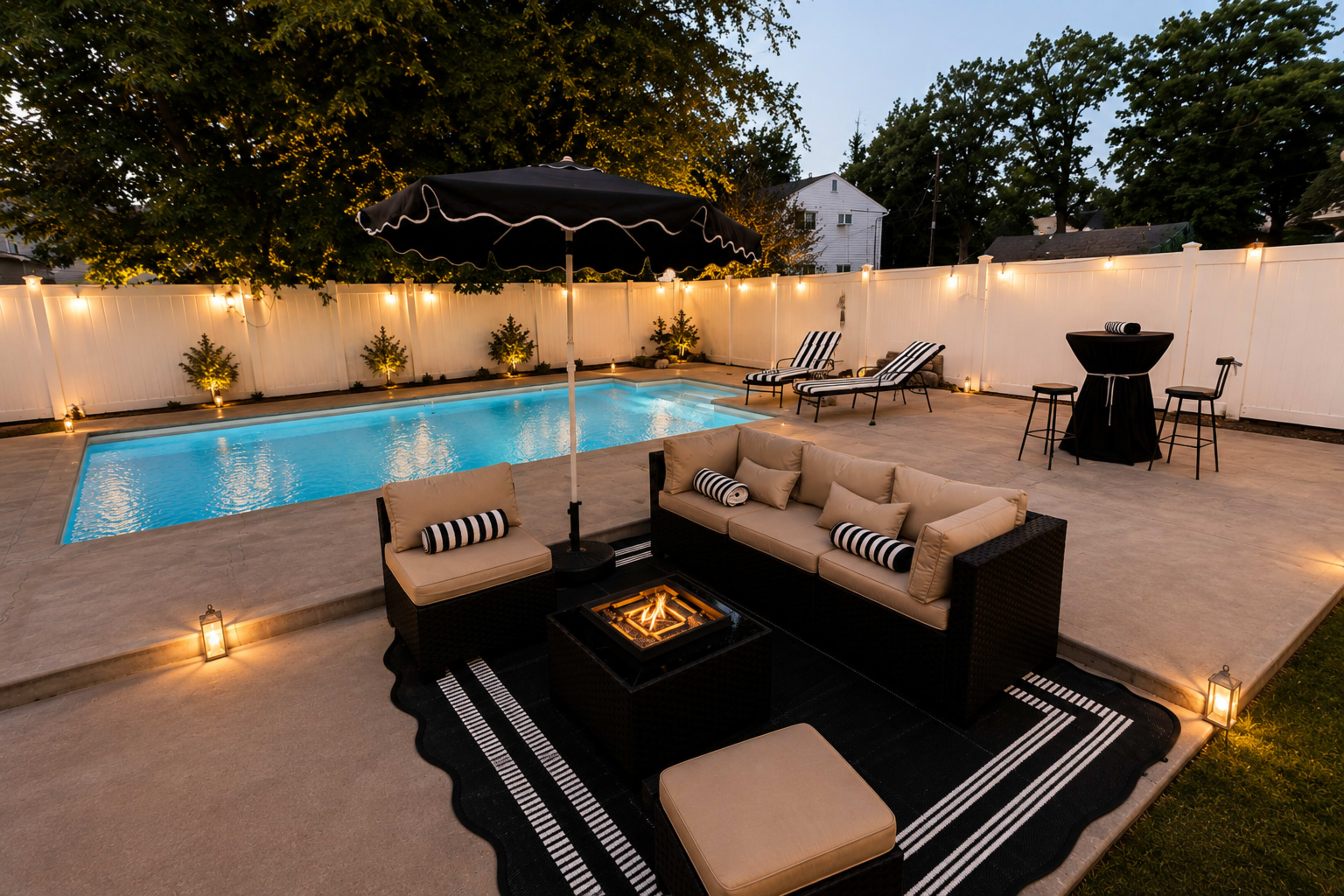 The image shows a backyard pool area featuring a swimming pool, lounge chairs, a black umbrella, a seating arrangement with a fire pit, and lighting along the perimeter.