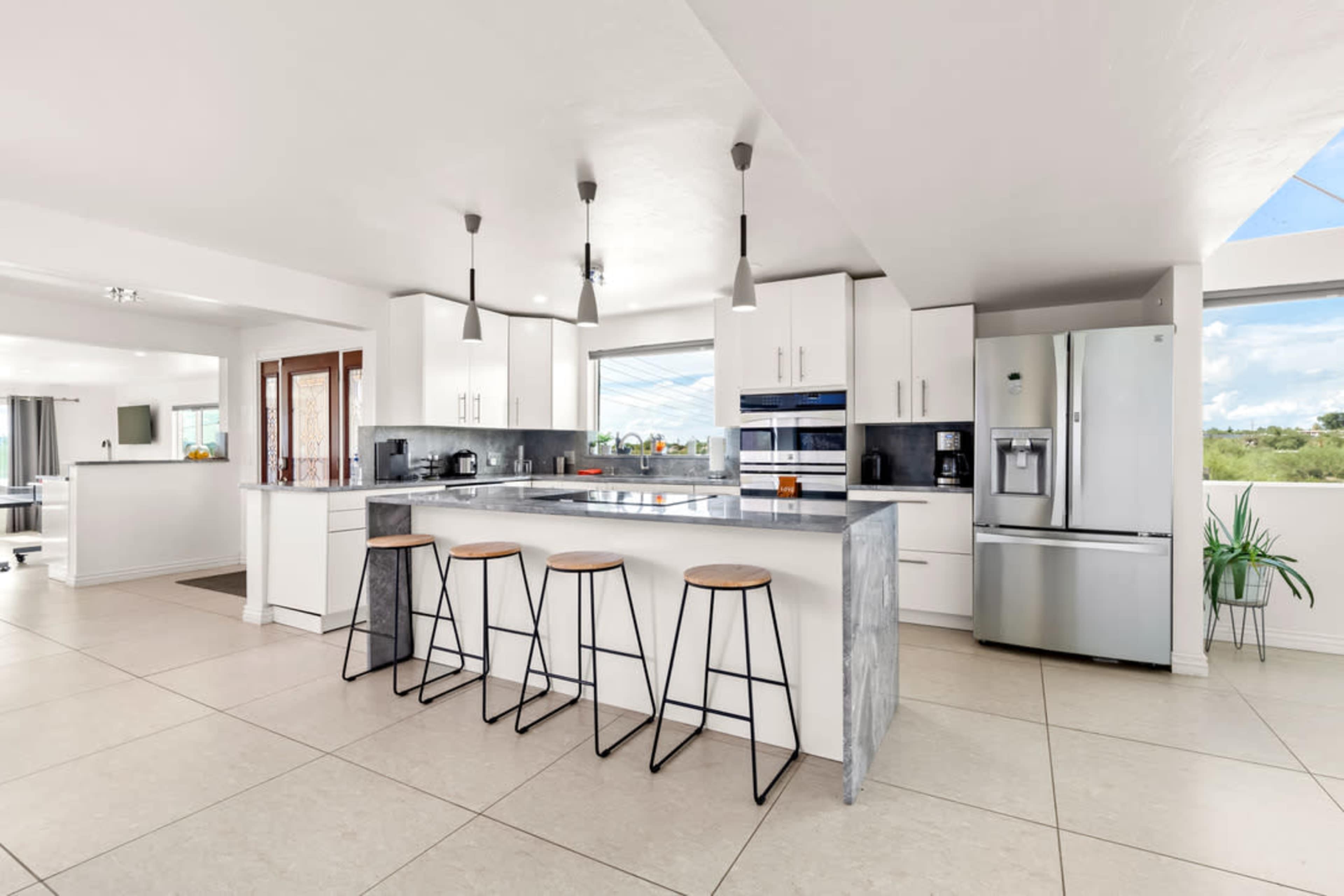 A modern kitchen features a central island with three stools, white cabinetry, stainless steel appliances, and large windows allowing natural light.