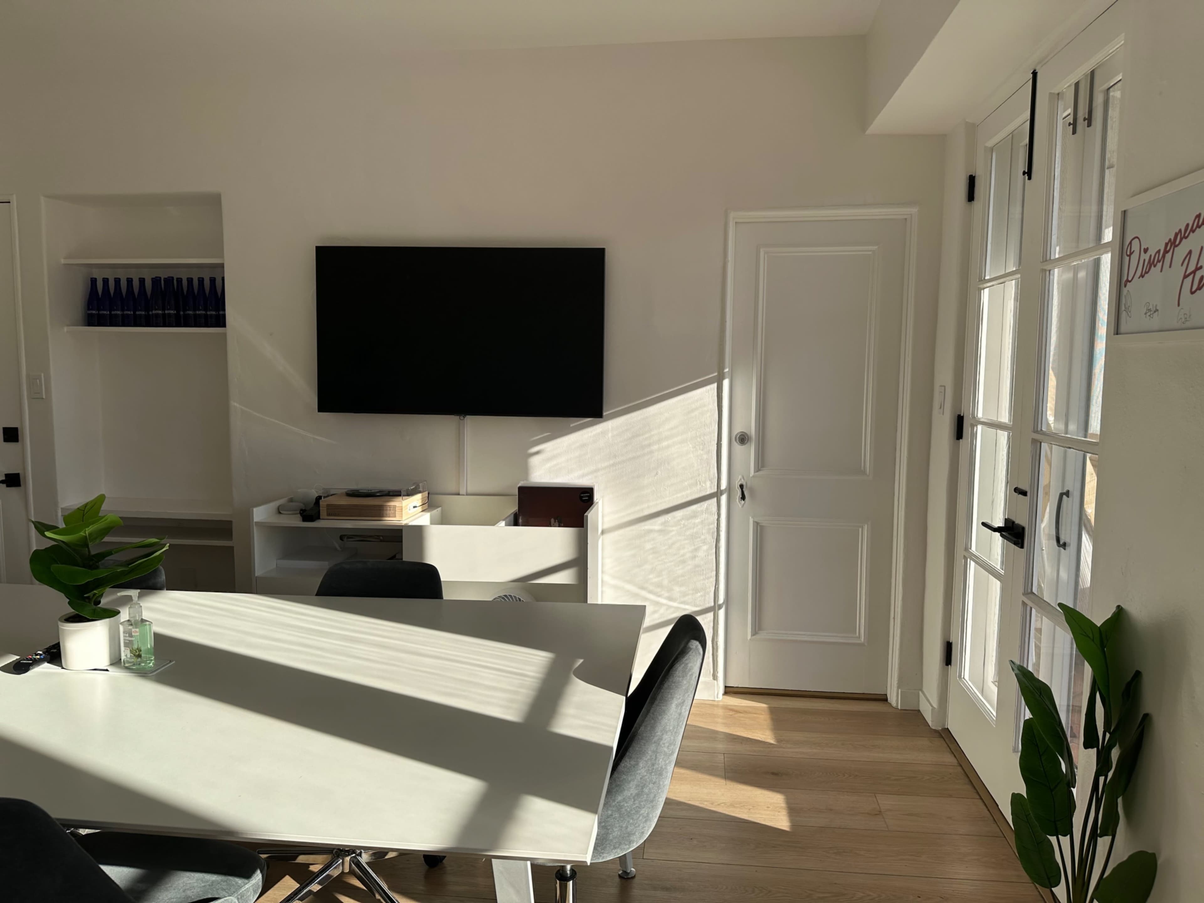 A modern living space features a wall-mounted television, a dining table with chairs, and light streaming through a door.