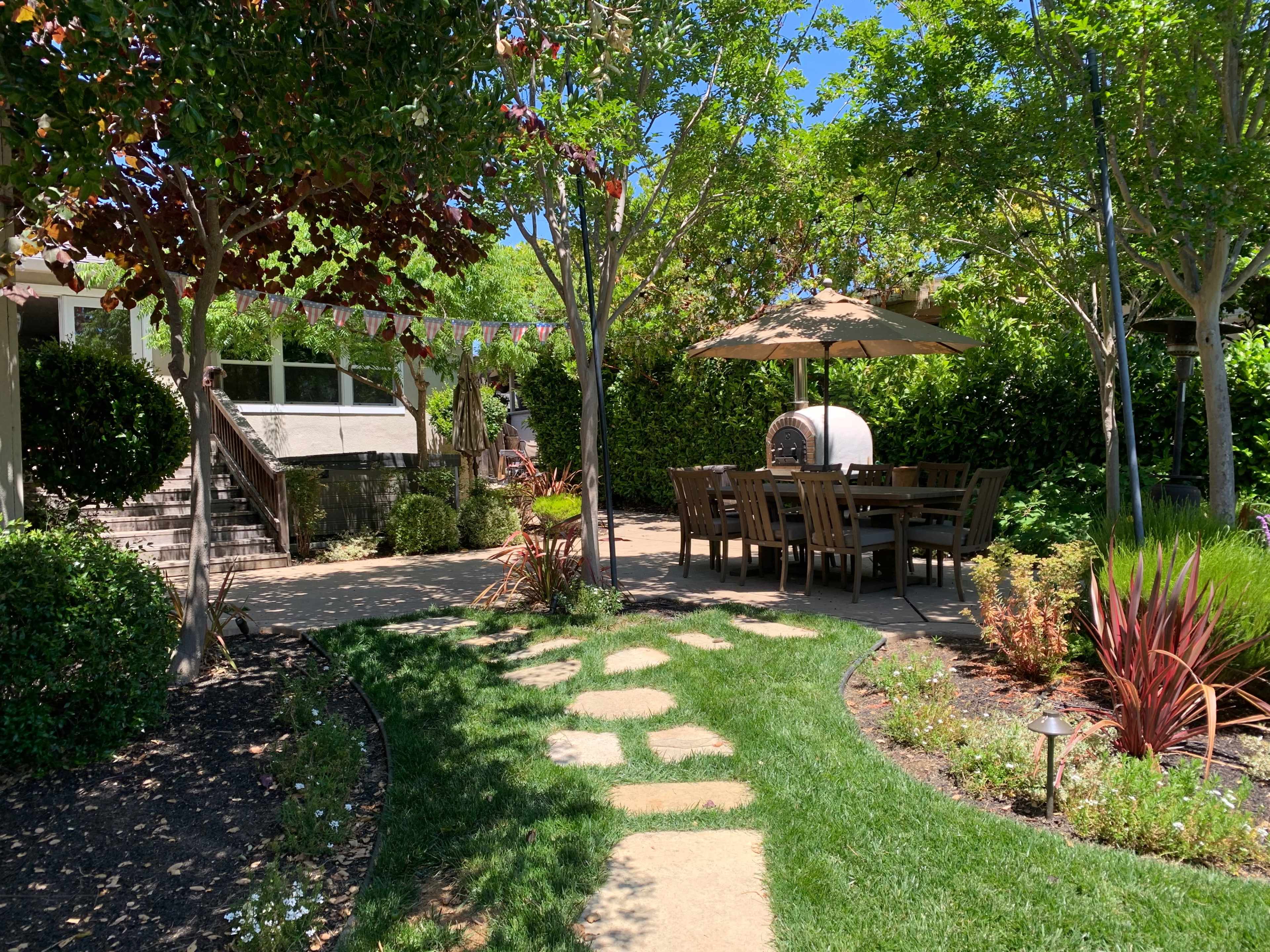 A landscaped backyard features a stone pathway leading to a dining area under a patio umbrella, surrounded by greenery and flowering plants.