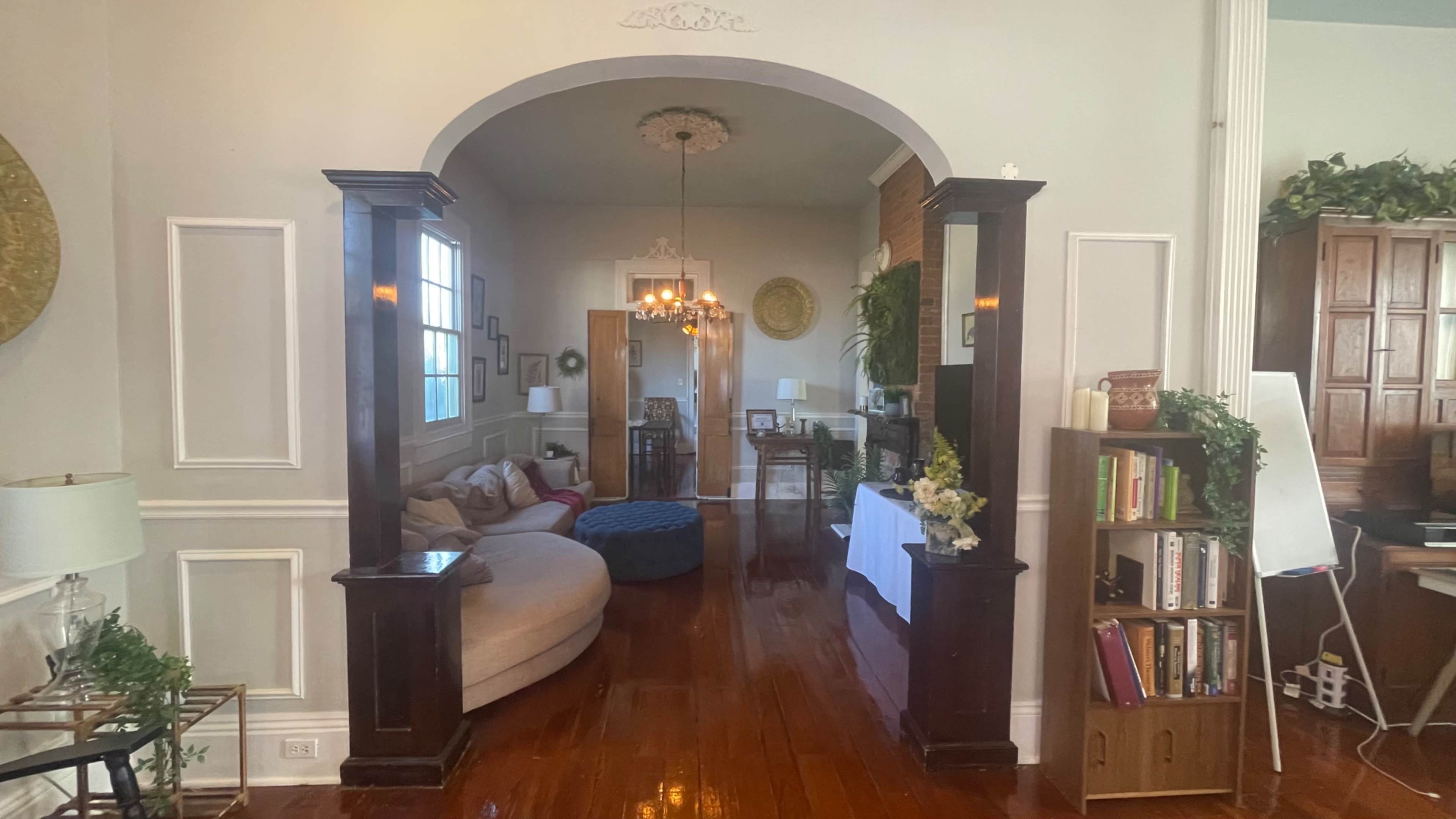 The image shows a spacious living area with an arched entryway, hardwood floors, and a blend of furniture including a large sofa, a round ottoman, and a bookshelf.