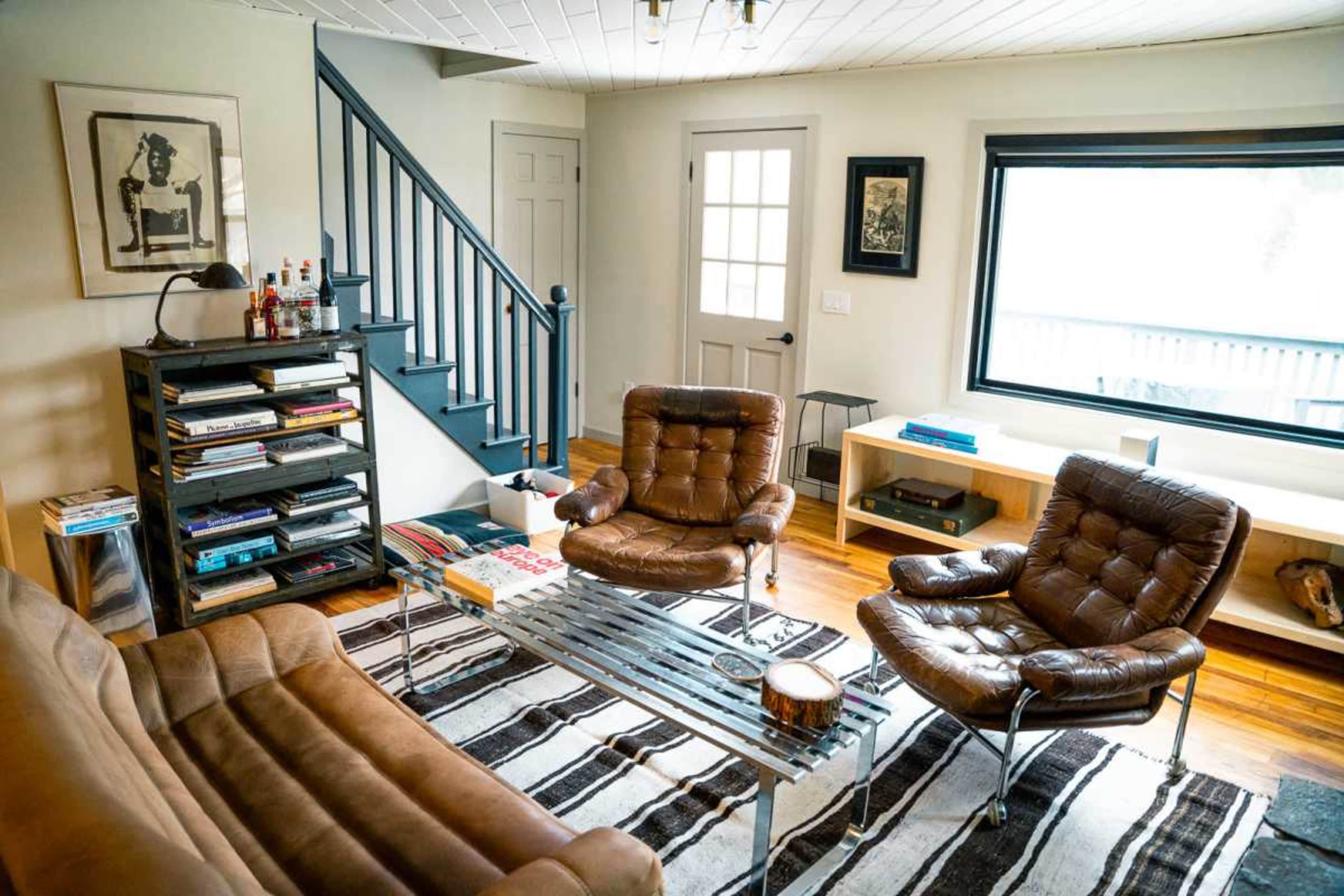 The image shows a living room featuring brown leather chairs and a sofa, a striped area rug, a glass coffee table, and a staircase leading to an upper level.