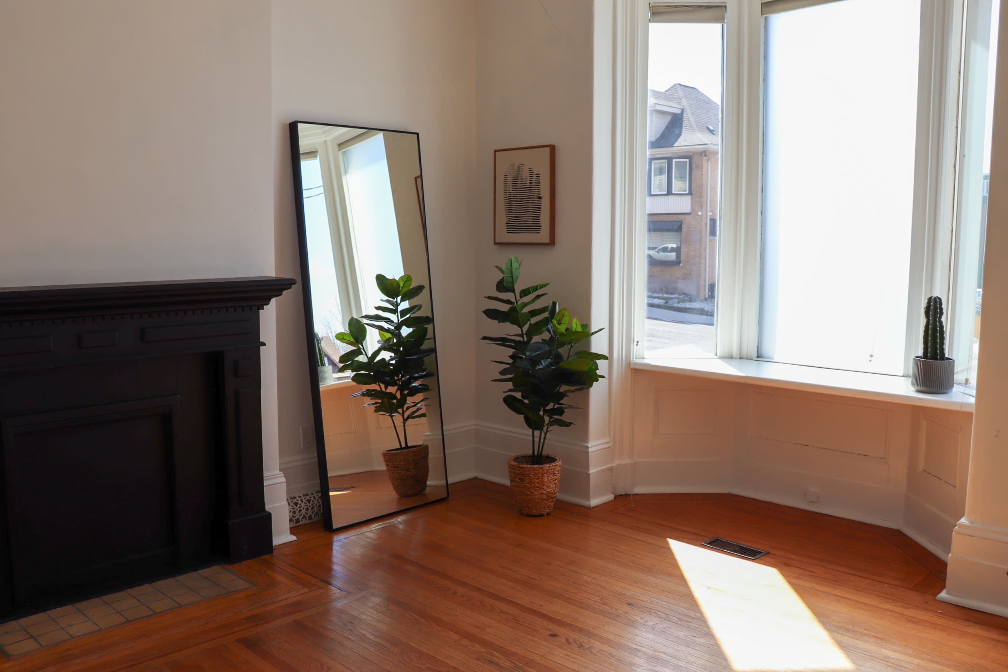 The image shows a bright room with a large mirror and two potted plants beside a bay window, featuring hardwood floors and a fireplace.