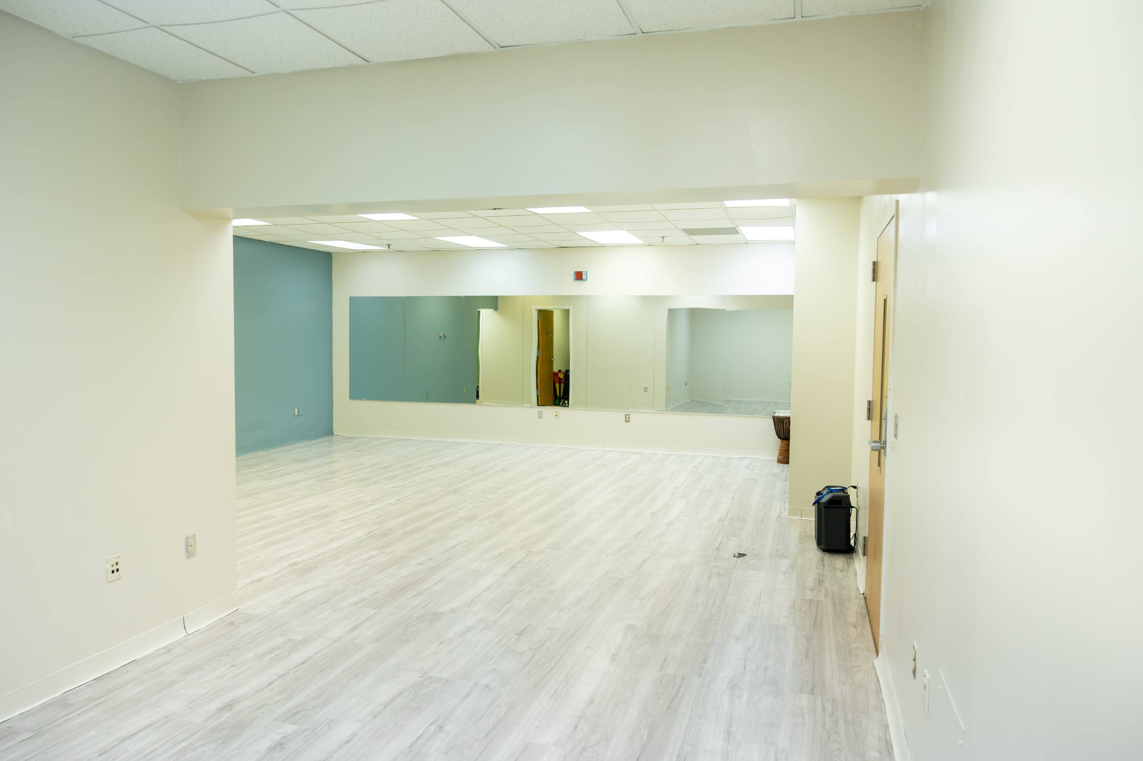 The image shows a brightly lit, empty room with light-colored flooring and large mirrors on one wall.
