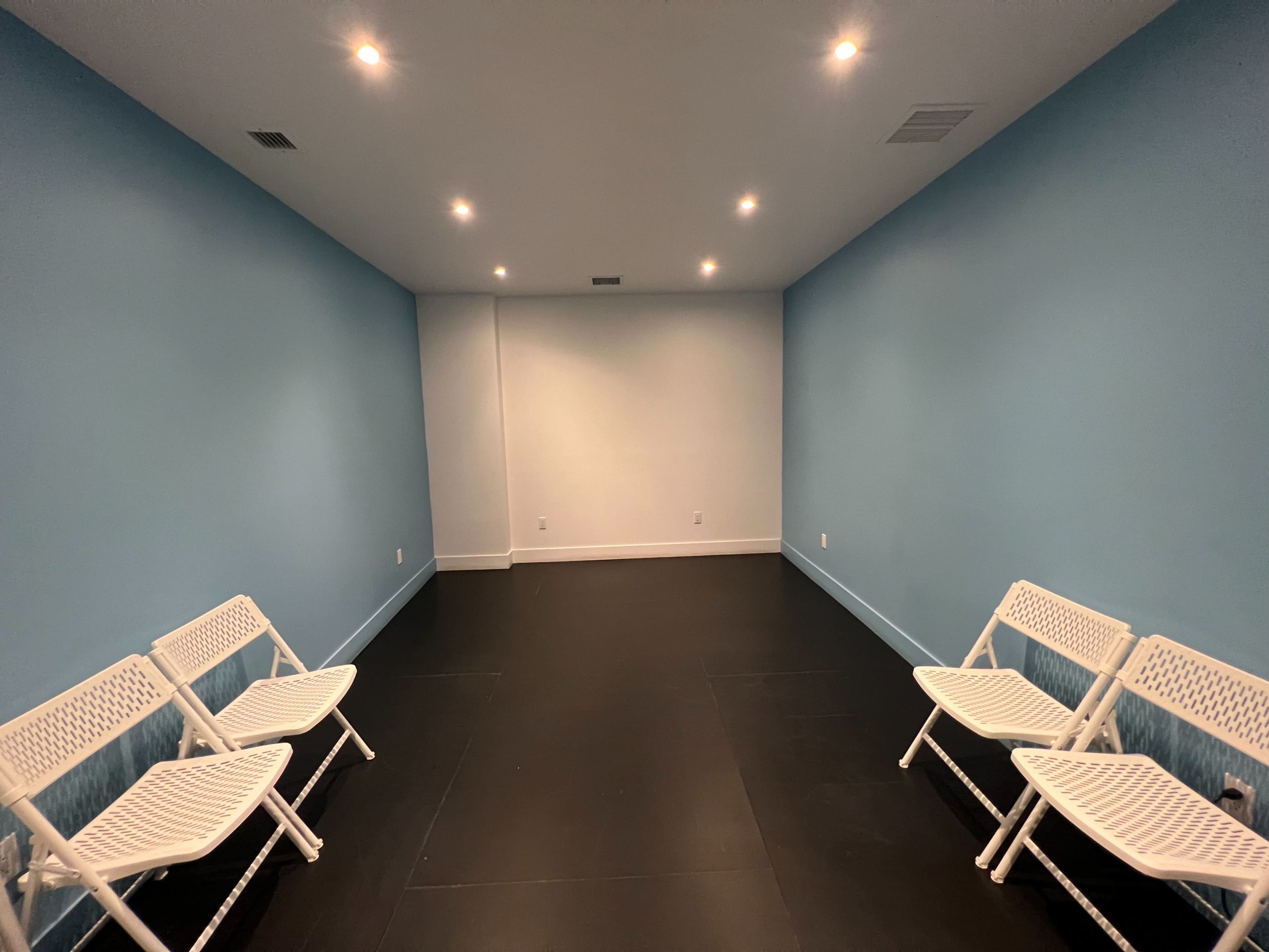 The image shows a sparsely furnished room with light blue walls, black flooring, and four white folding chairs arranged in pairs along one side.