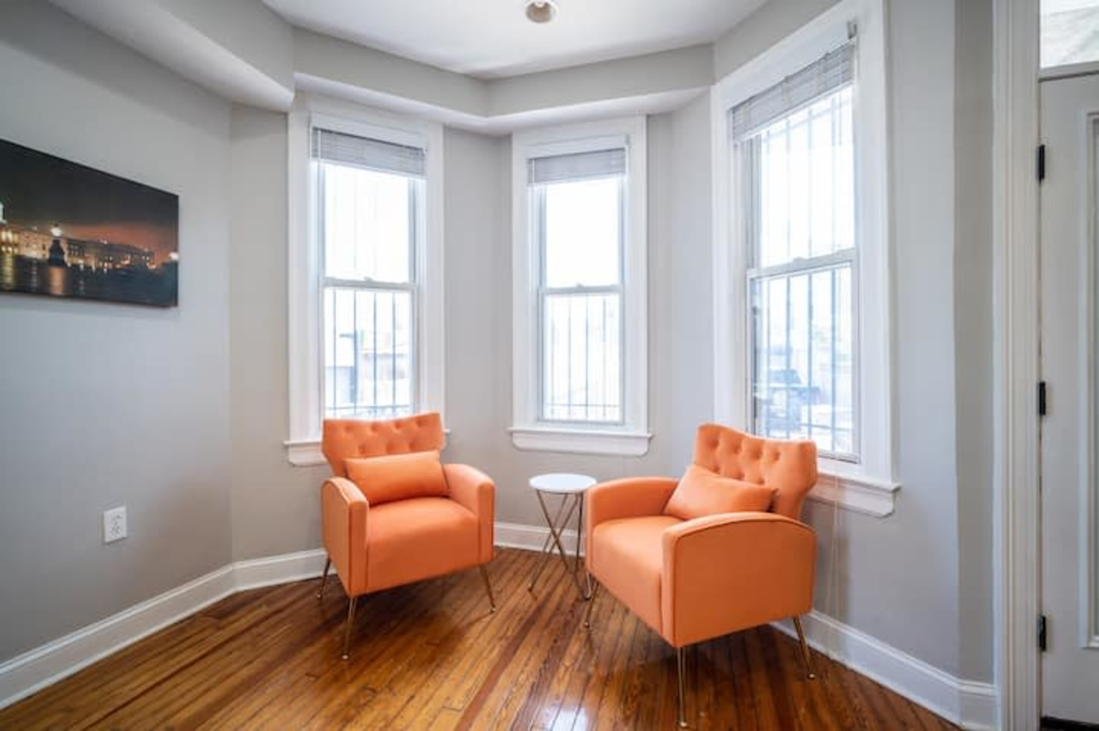 The image shows a cozy corner with two orange armchairs and a small table, illuminated by natural light from the windows.