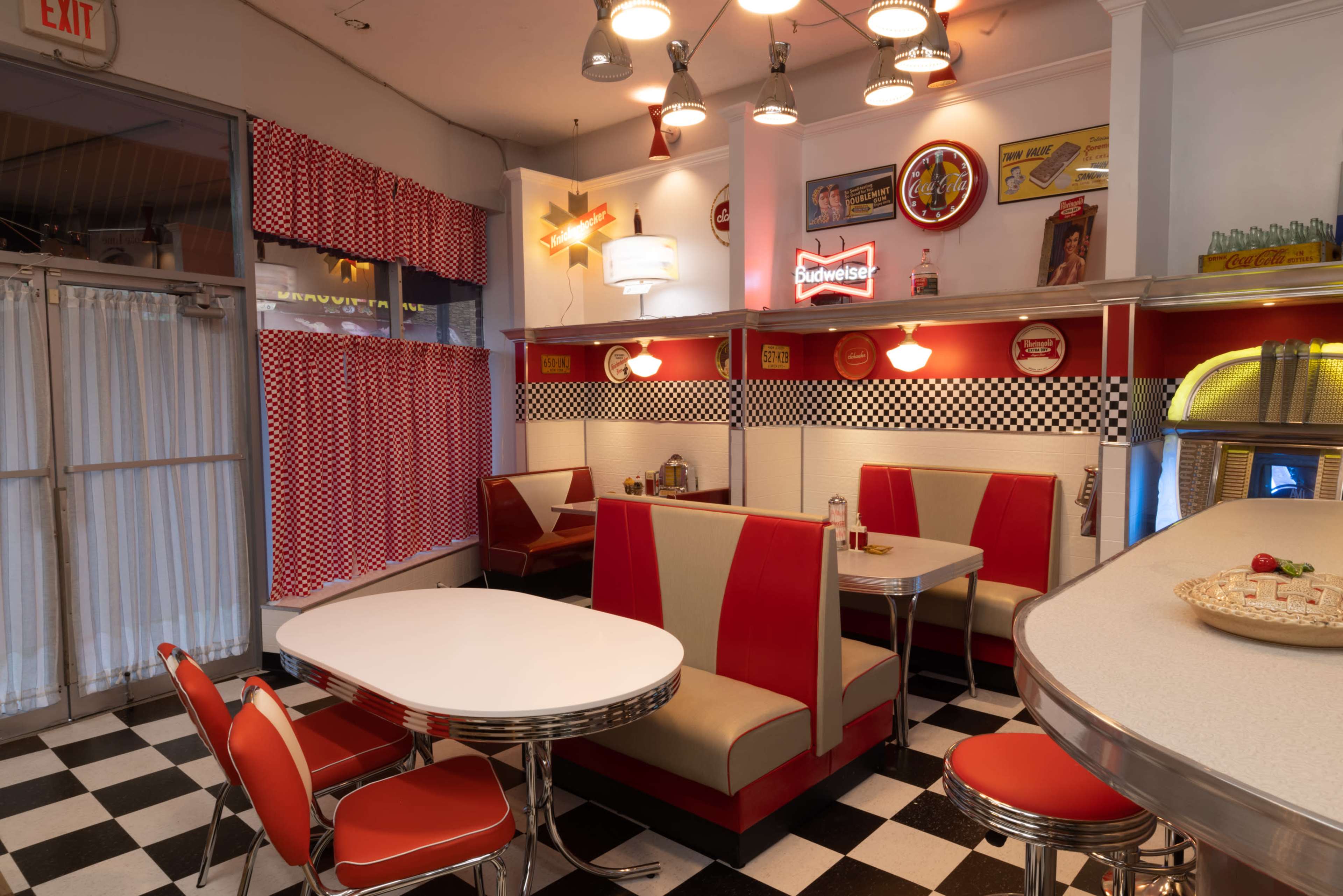 The interior of a retro-style diner with checkered floors, red and white decor, and booth seating arranged around a central table.