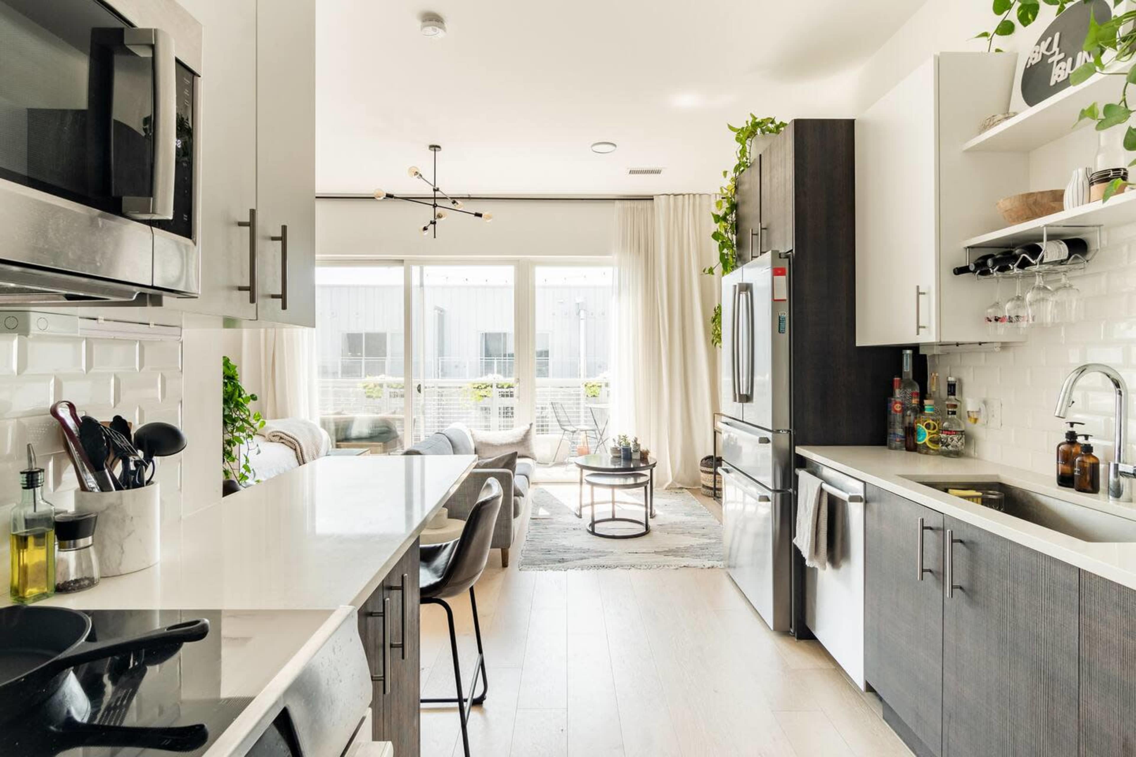 A modern kitchen with stainless steel appliances and a white countertop opens into a cozy living area adorned with plants and natural light.