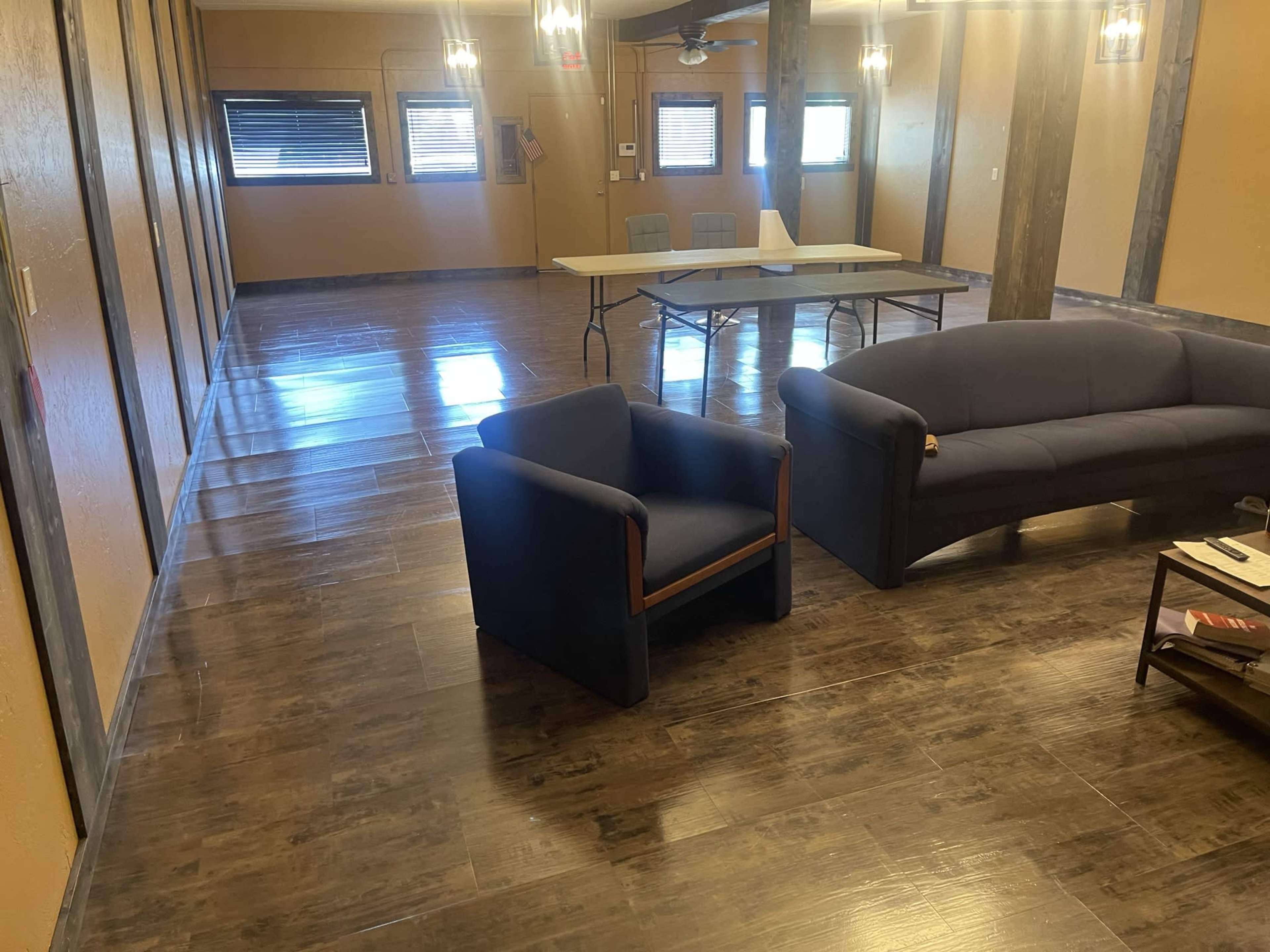 A spacious room with polished brown flooring, a blue couch, a blue armchair, and a table set up in the center.