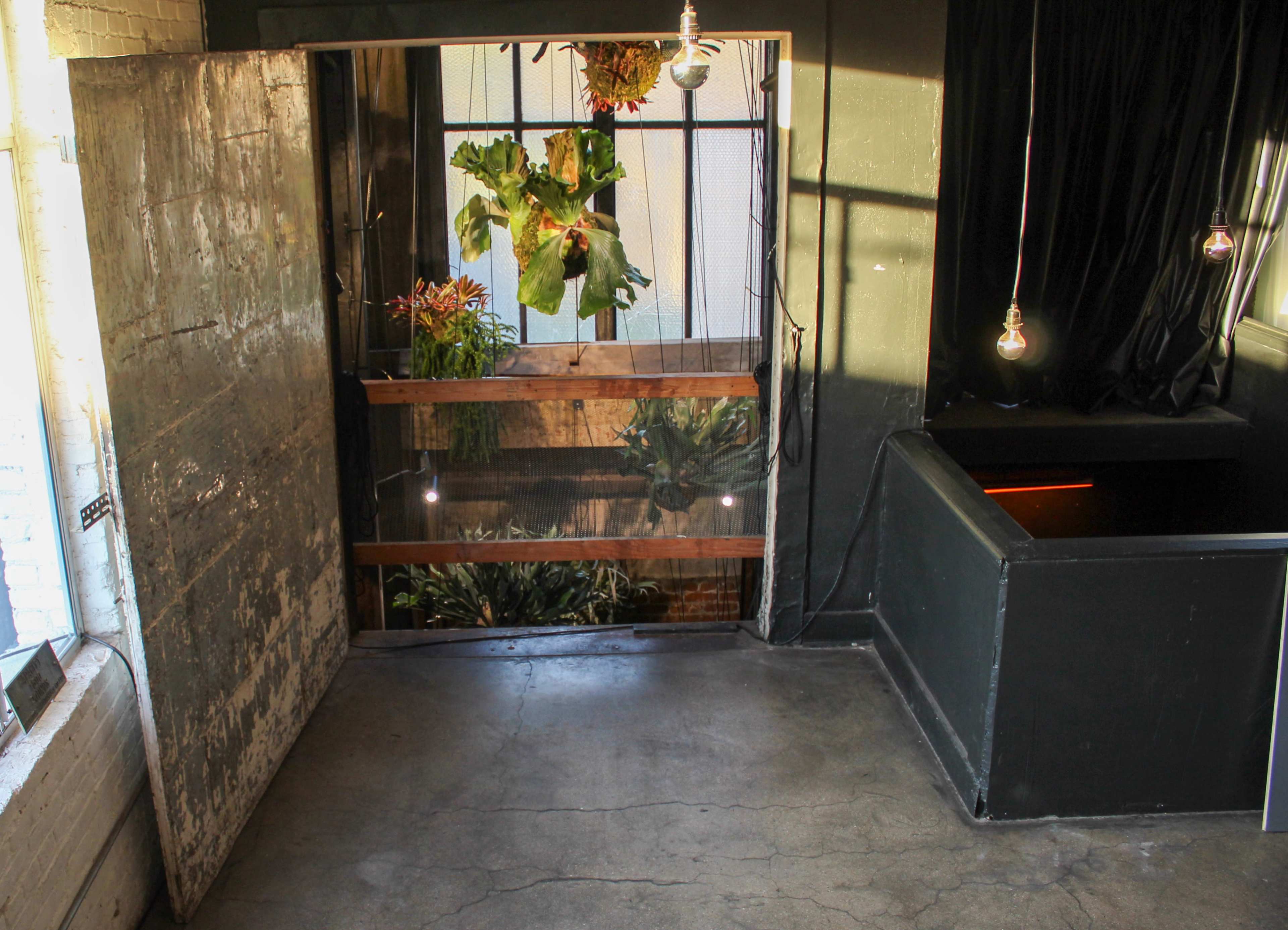 The image shows a room with a partially open metal door revealing a view of greenery outside, along with a dark wall and a small black counter in the corner.