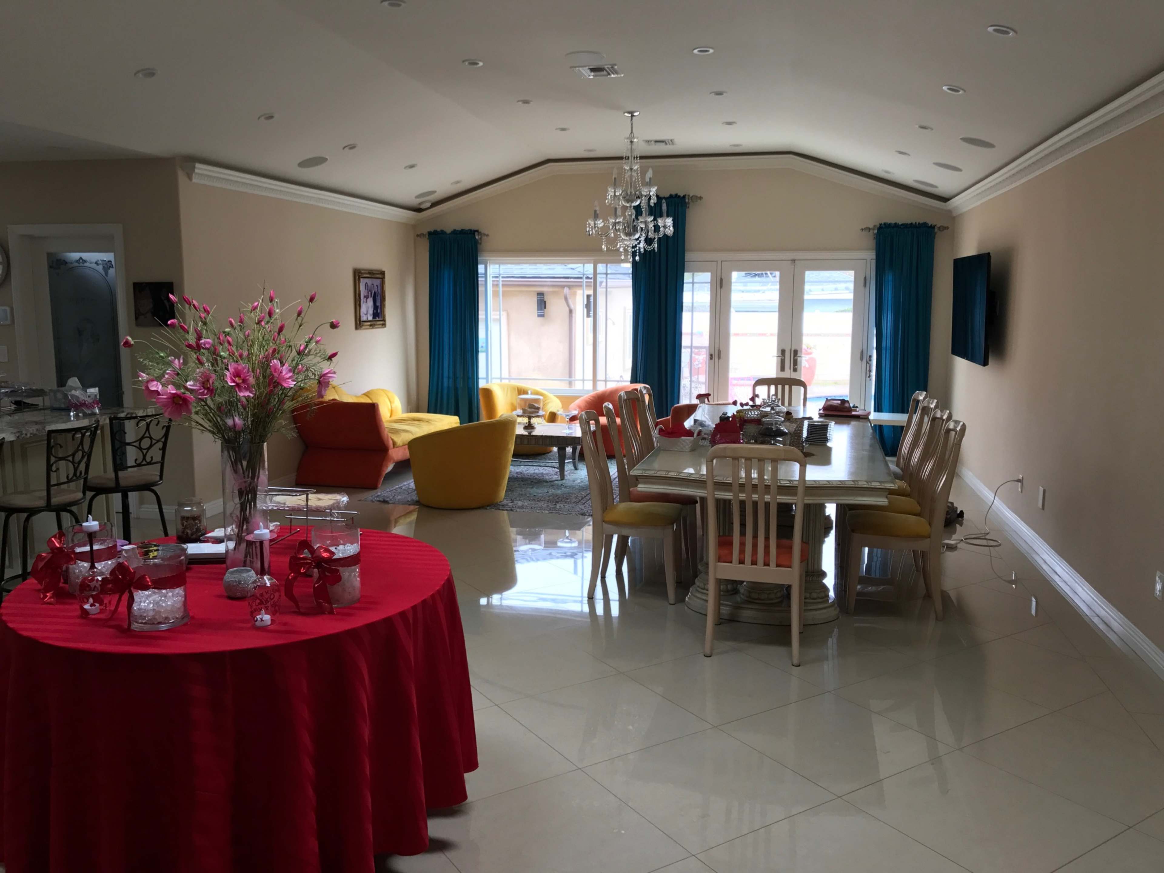A bright living and dining area features a large table with red decorations, colorful chairs, and large windows allowing natural light to enter.