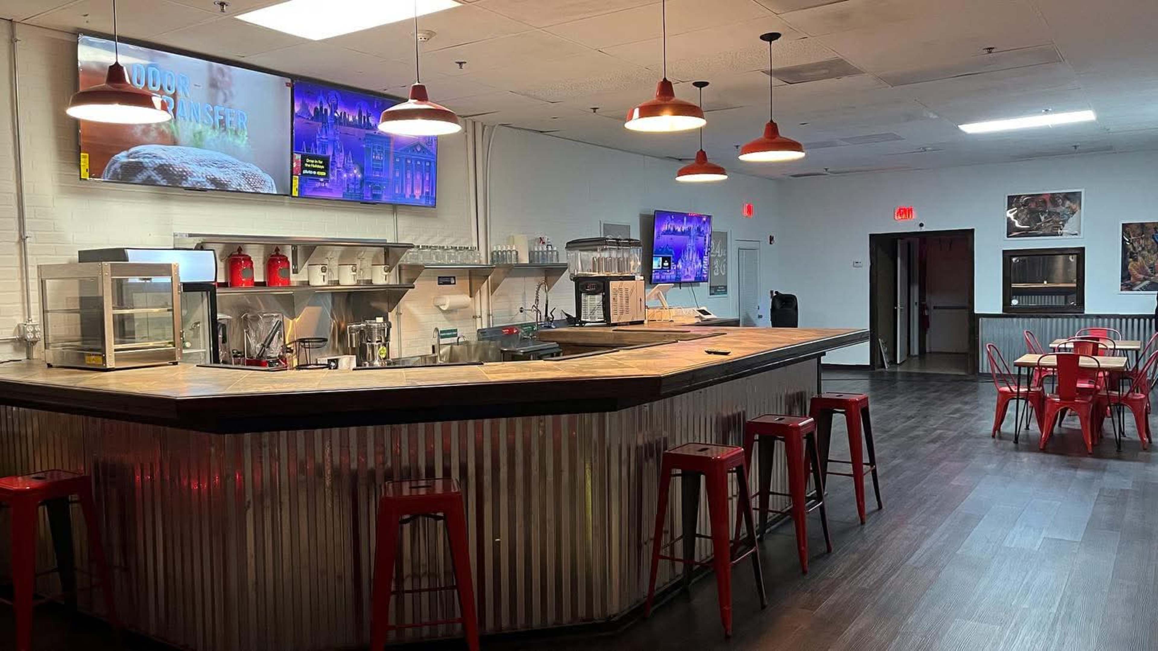 The image shows a modern café or bar interior with a wooden counter, red stools, and monitors displaying information on the walls.