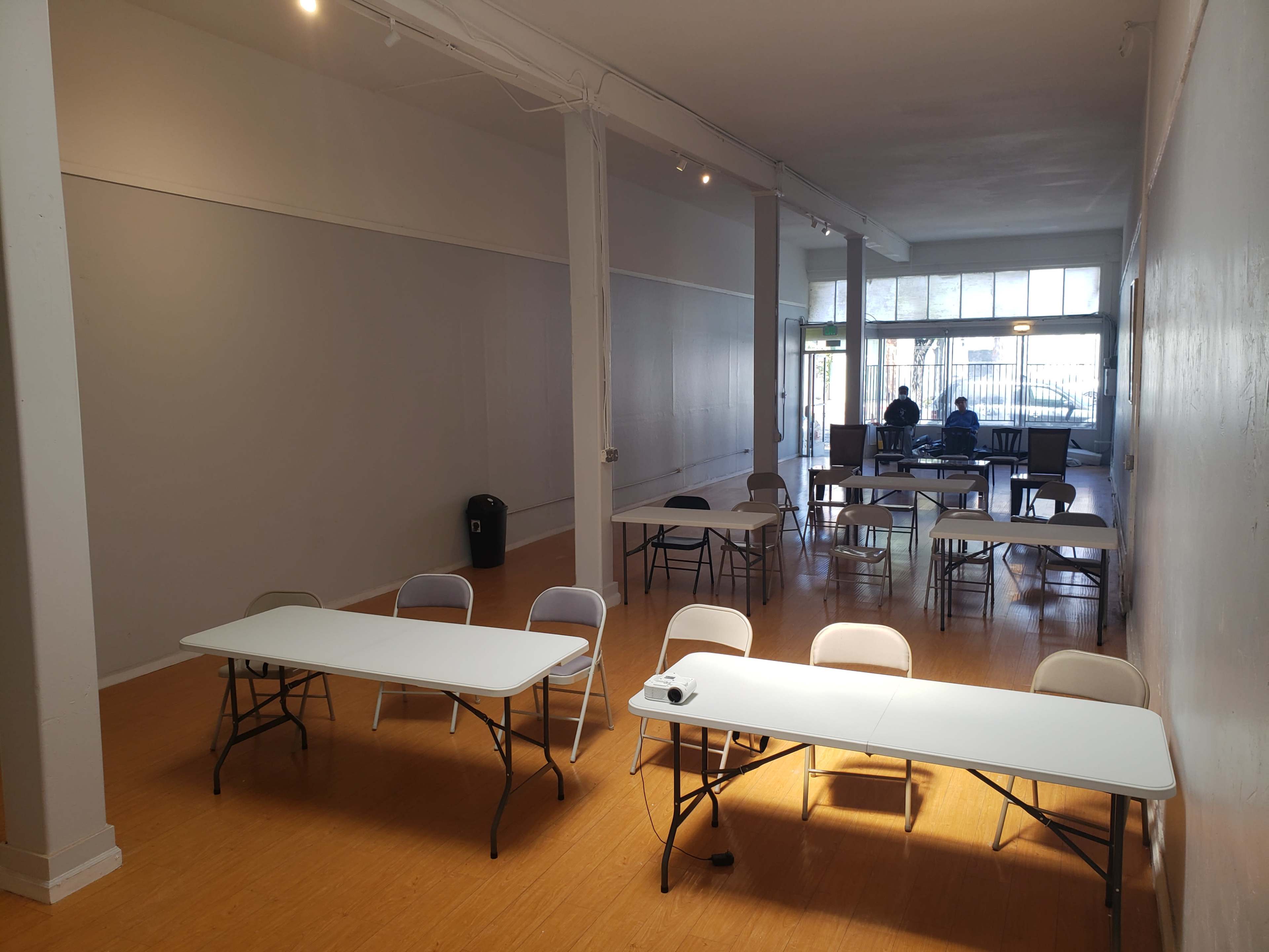 An empty room with several folding tables and chairs arranged, featuring two people seated at a table in the background.